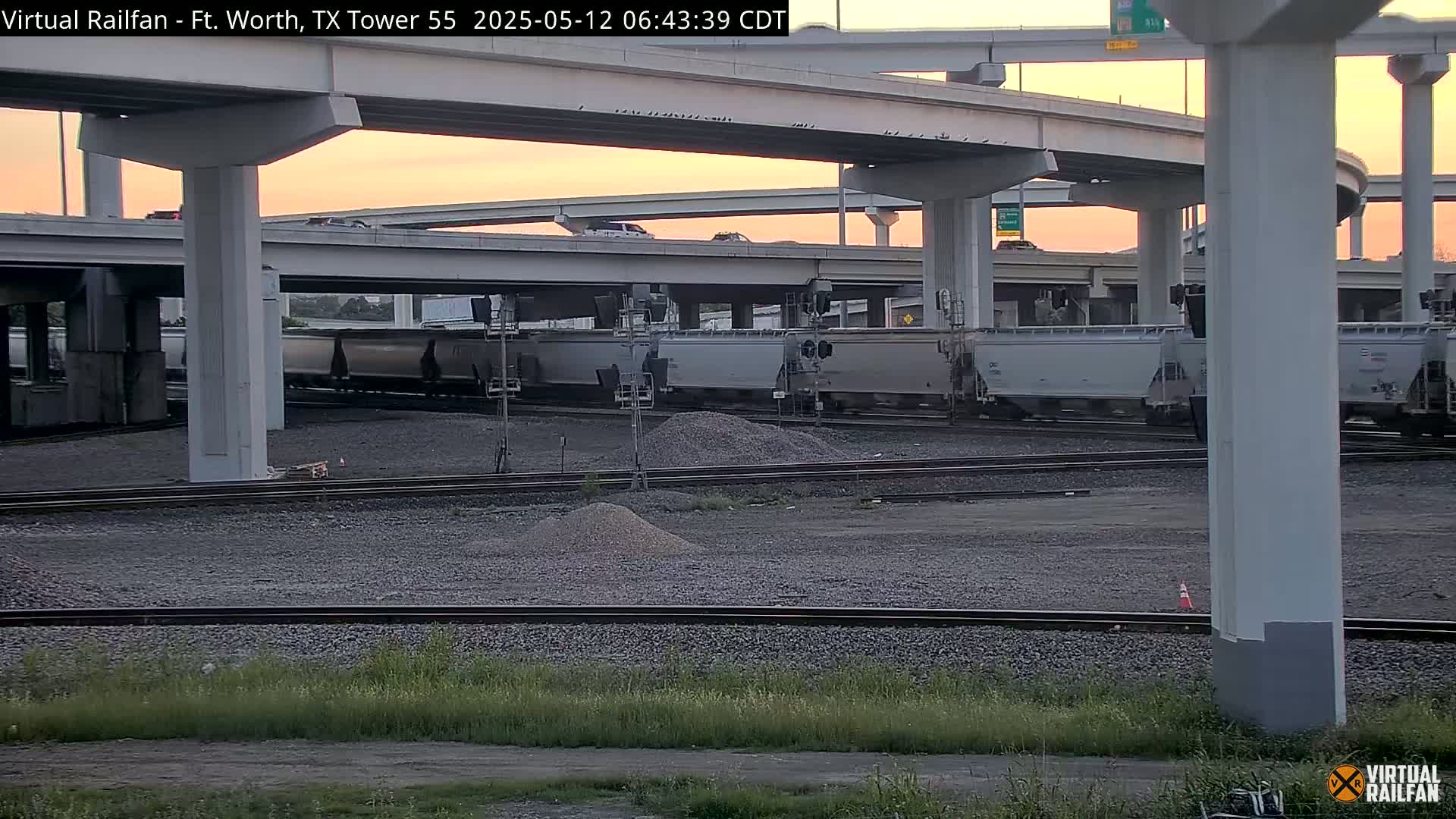 A freight train sits on railroad tracks below an elevated highway during a dusky sunrise.
