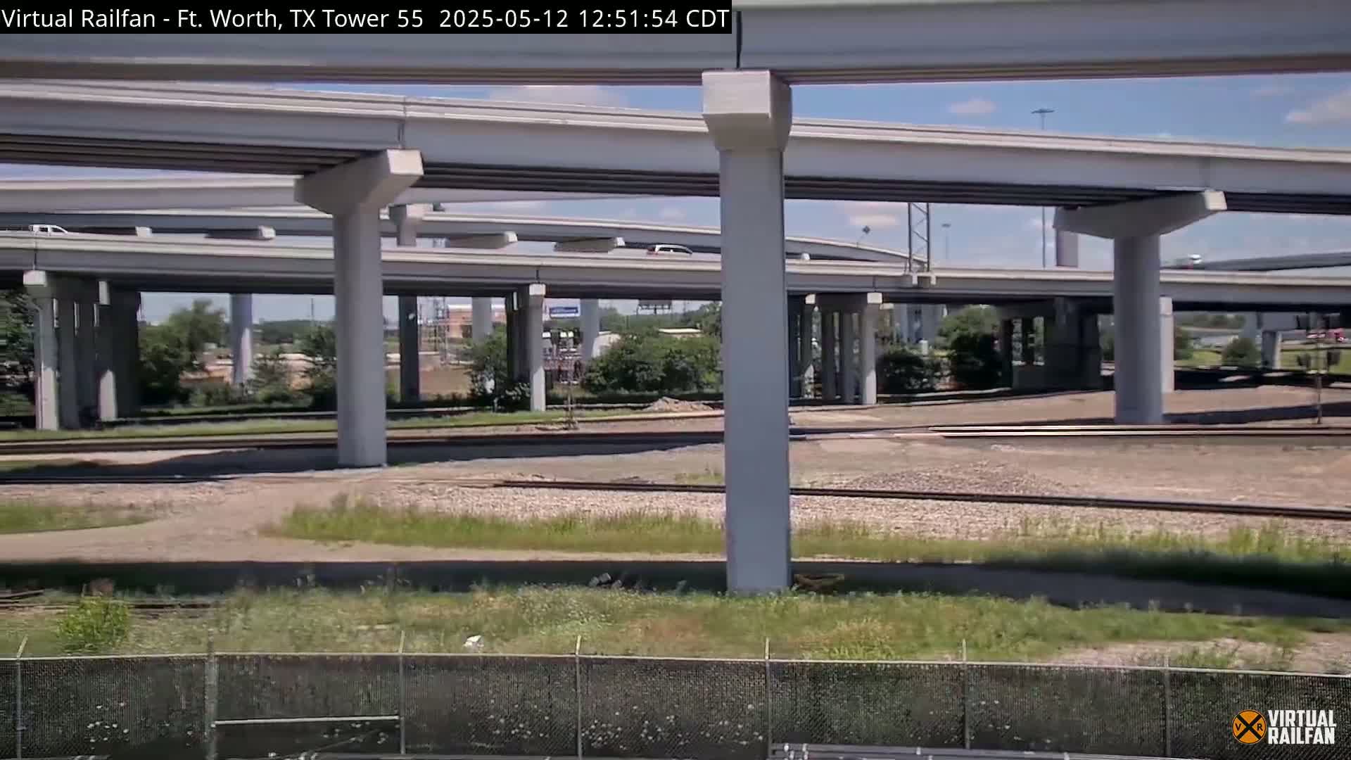 A highway overpass with multiple levels and supports sits above a set of railroad tracks on a sunny day.