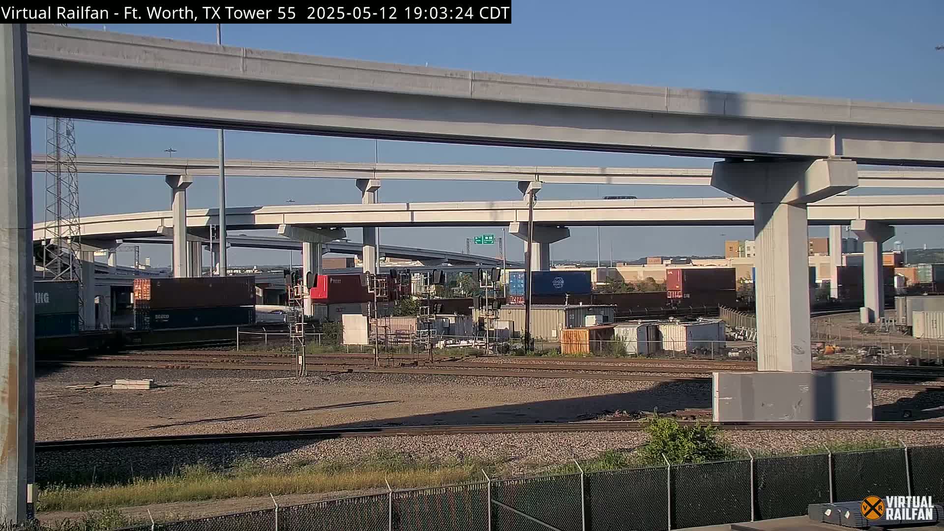 A freight train sits below an elevated highway interchange on a sunny day.