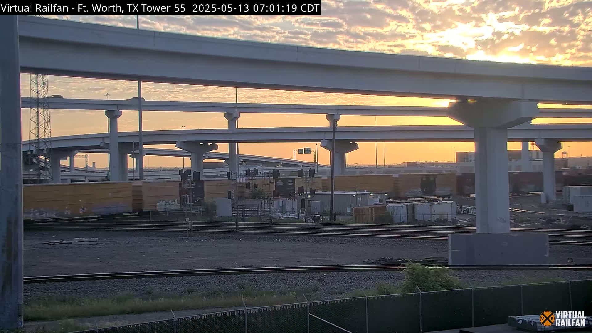 Railroad tracks and freight cars sit beneath a multi-level highway overpass during a sunrise with a partly cloudy sky.