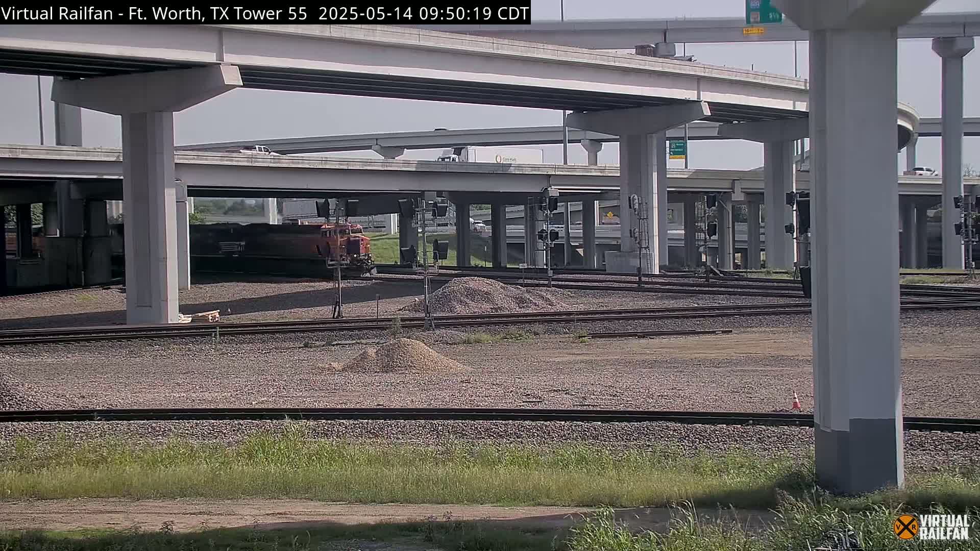 A freight train sits beneath a highway overpass on a sunny day, with gravel and railroad tracks in the foreground.