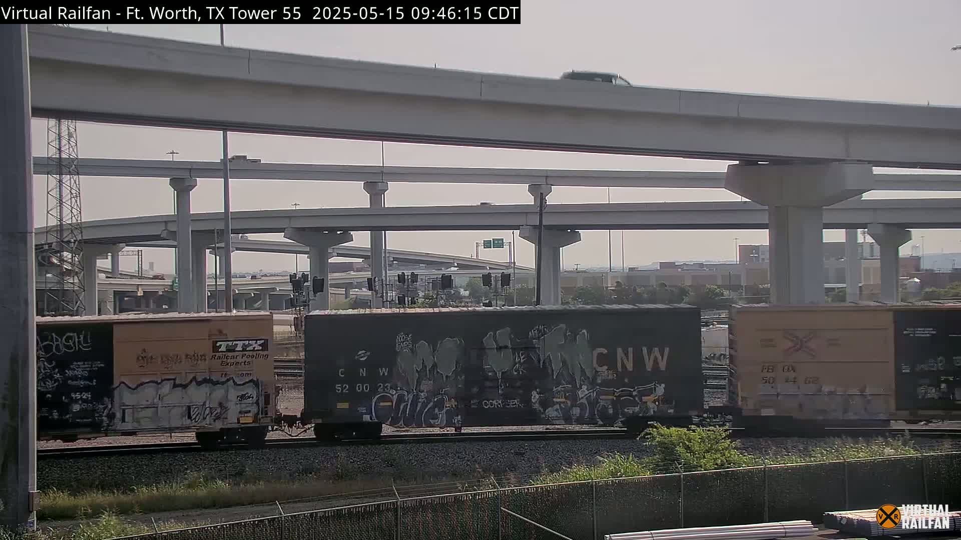 Under a hazy sky, a freight train with graffiti-covered cars sits beneath a multi-level highway overpass.