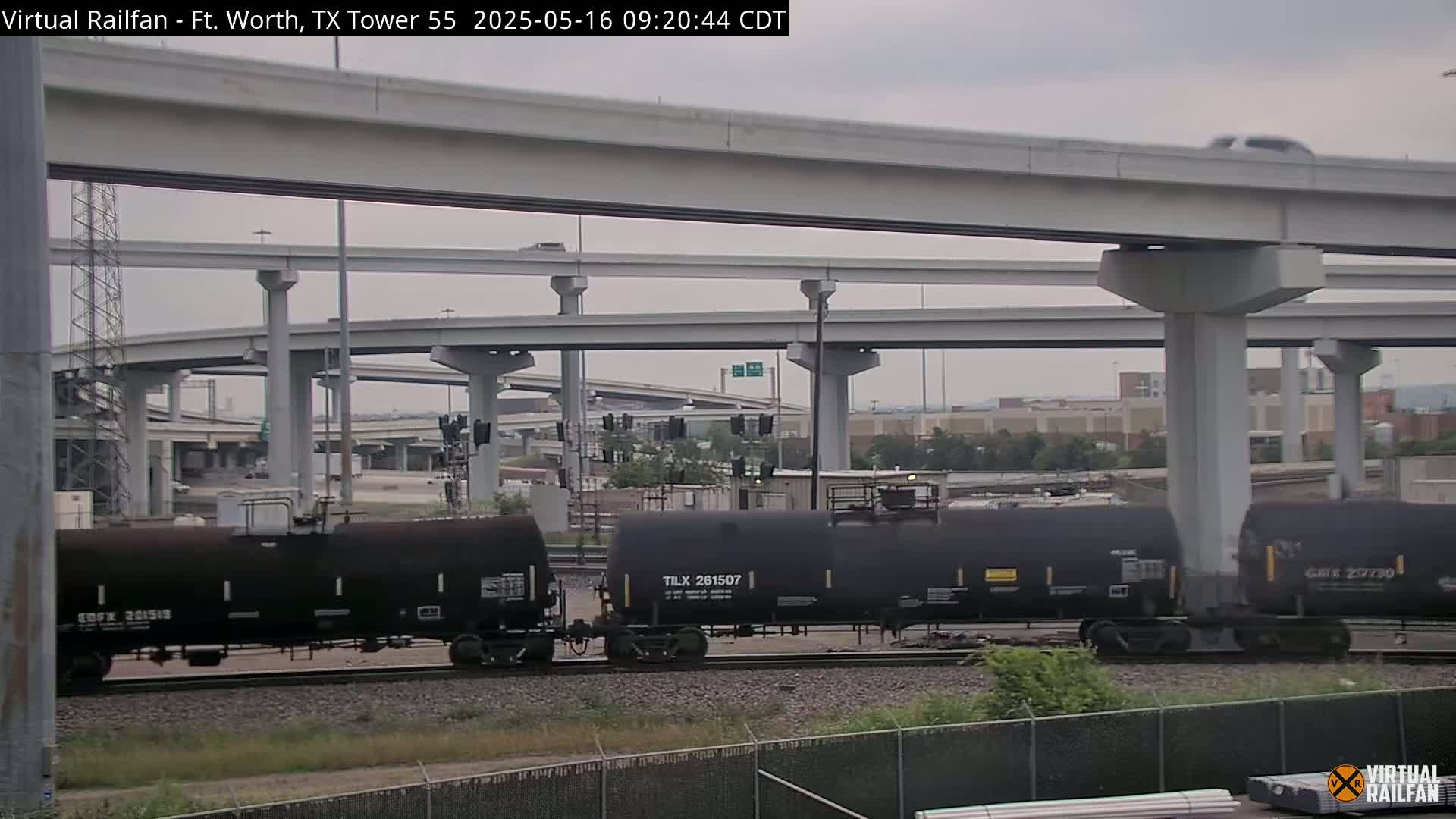 A freight train carrying tanker cars is visible below a complex overpass system on an overcast day.