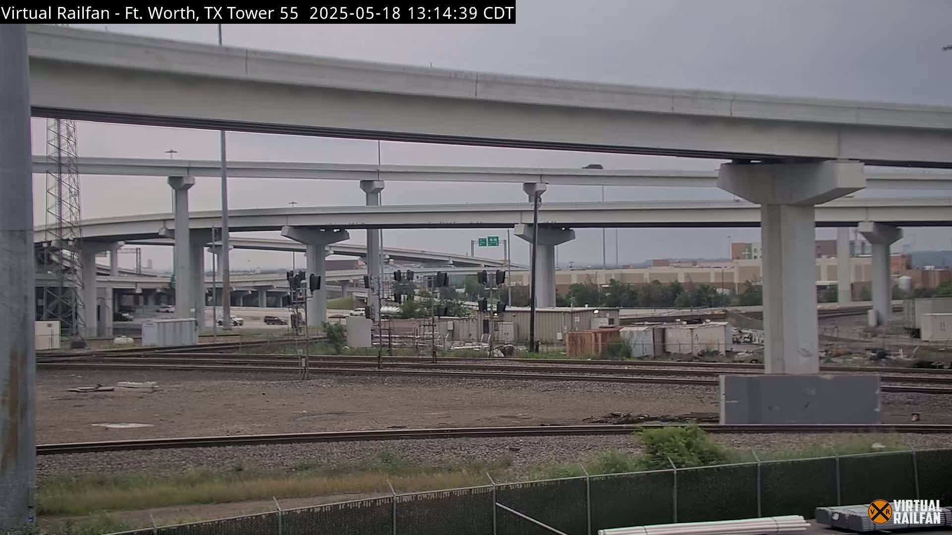 Under an overcast sky, a complex network of elevated highway overpasses and railroad tracks are seen, with some industrial buildings and vegetation in the background.