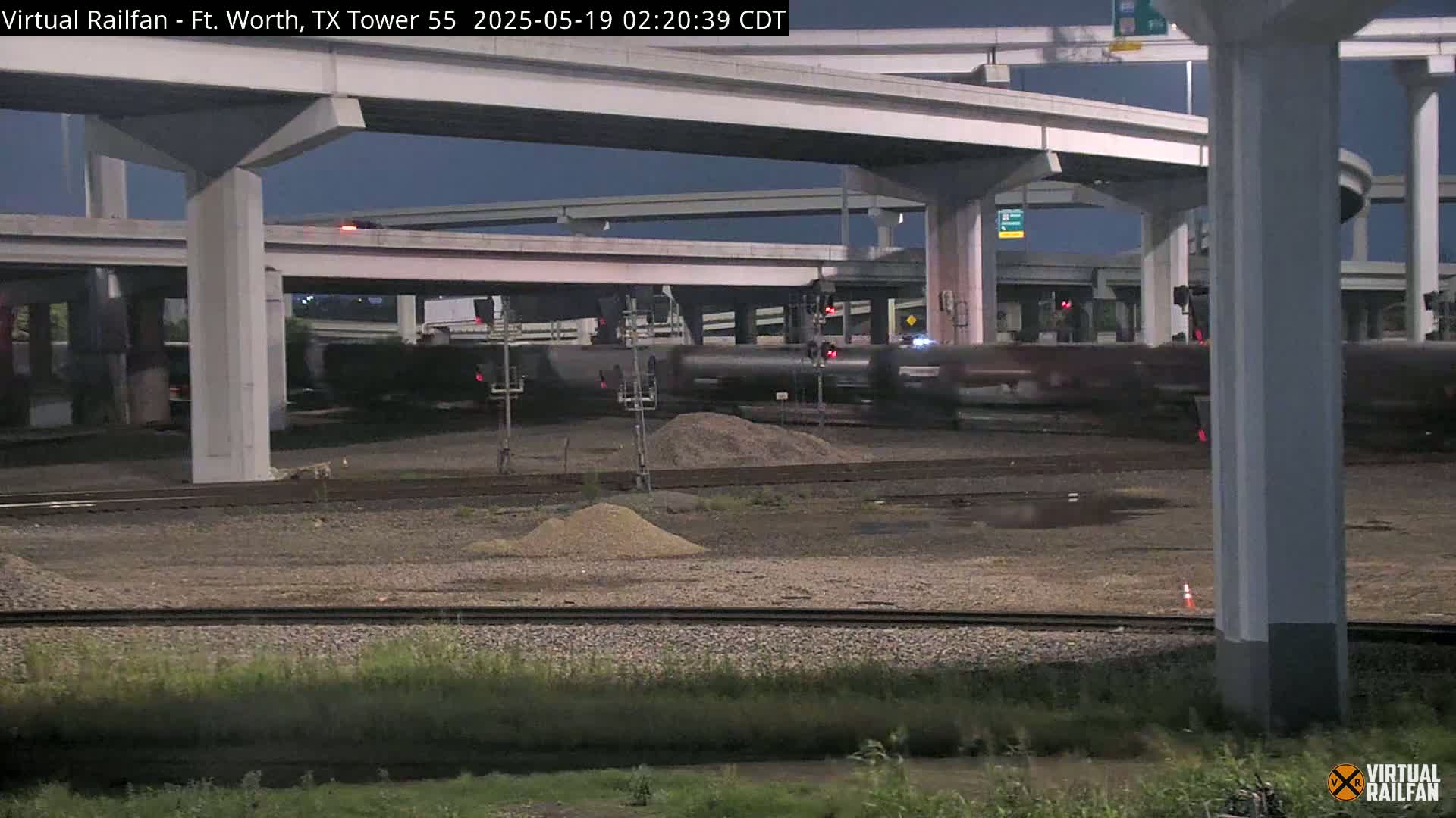 A freight train passes under a large highway overpass at night.