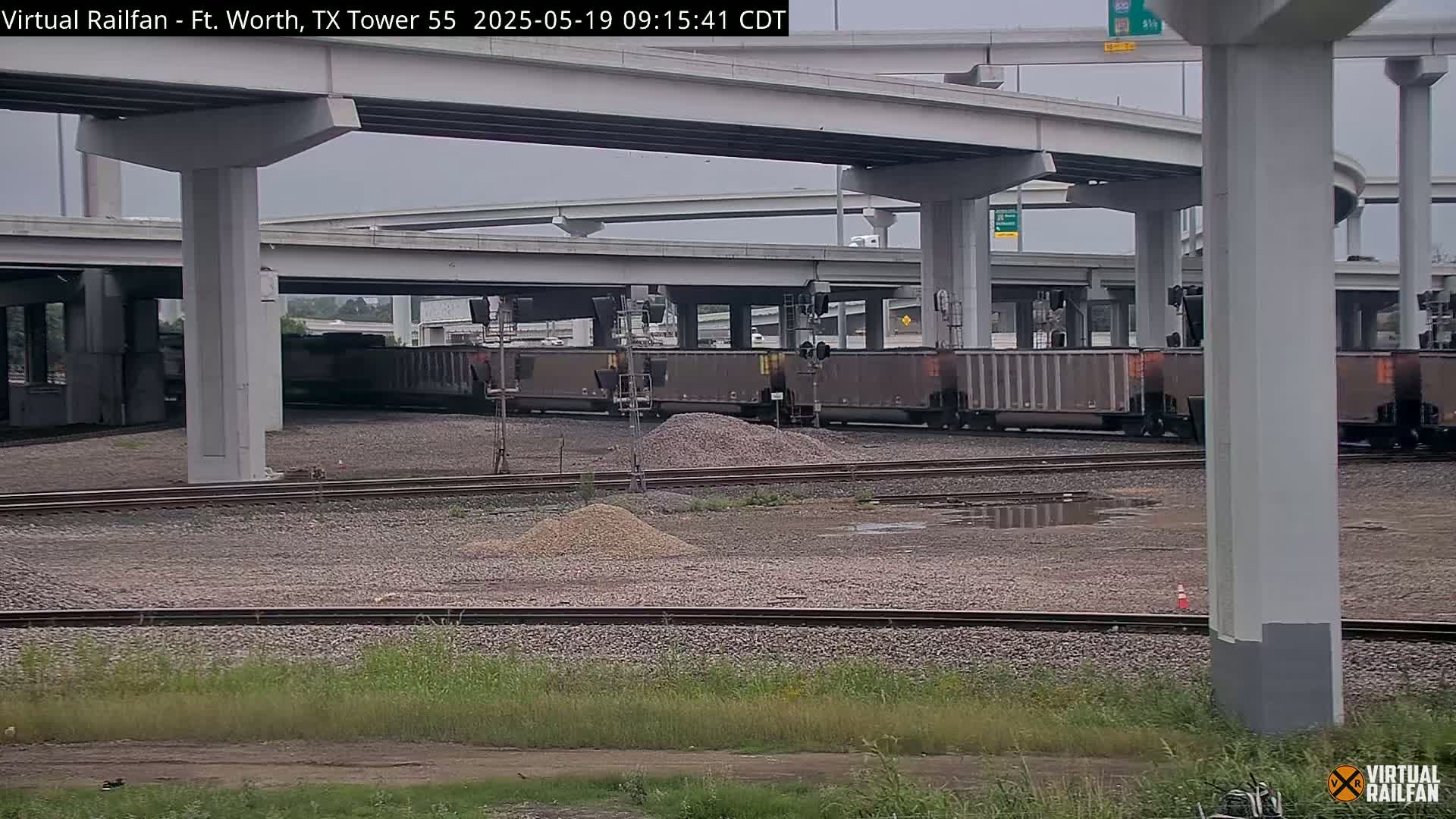 A freight train sits under a highway overpass on a partly cloudy day, with piles of gravel scattered nearby on a gravel rail yard.