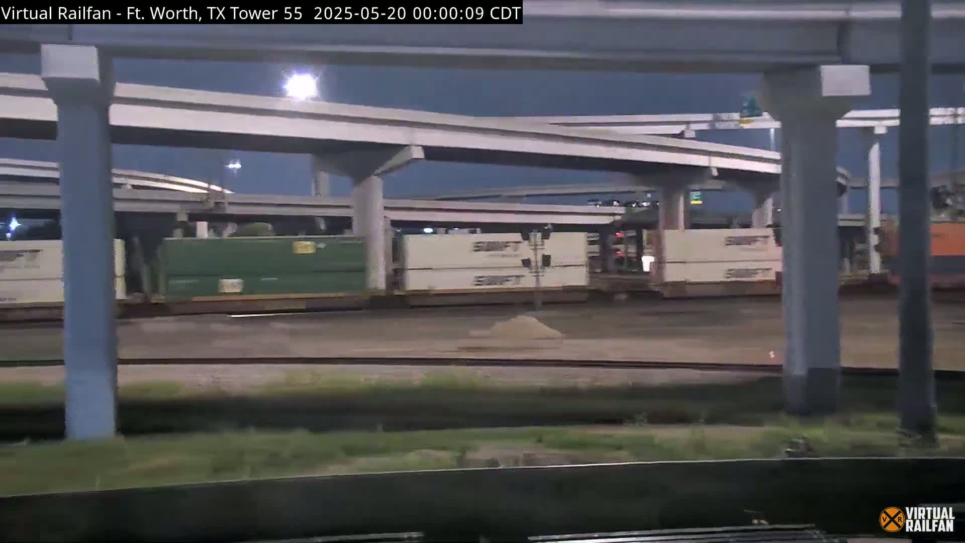 Under a dark night sky, a freight train with several boxcars sits near an elevated highway overpass.