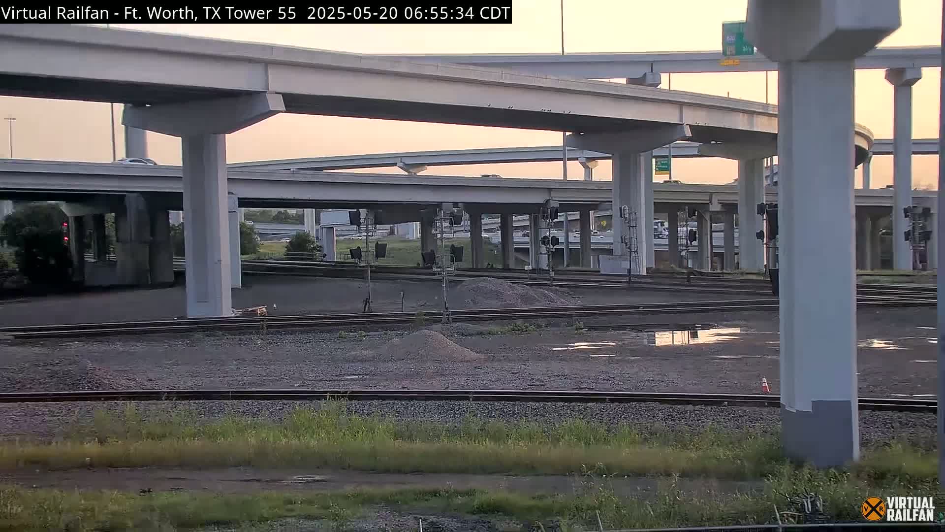 A view of a railway track and a large highway interchange at sunset, with some puddles of water on the ground.