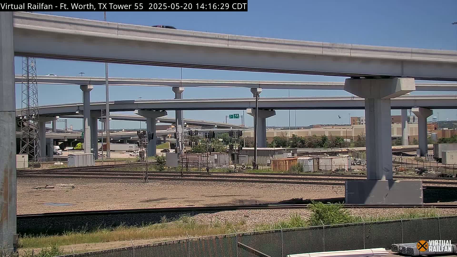 Under a clear blue sky, an elevated highway overpass and intersecting railway tracks are visible, with various industrial buildings and containers in the background.