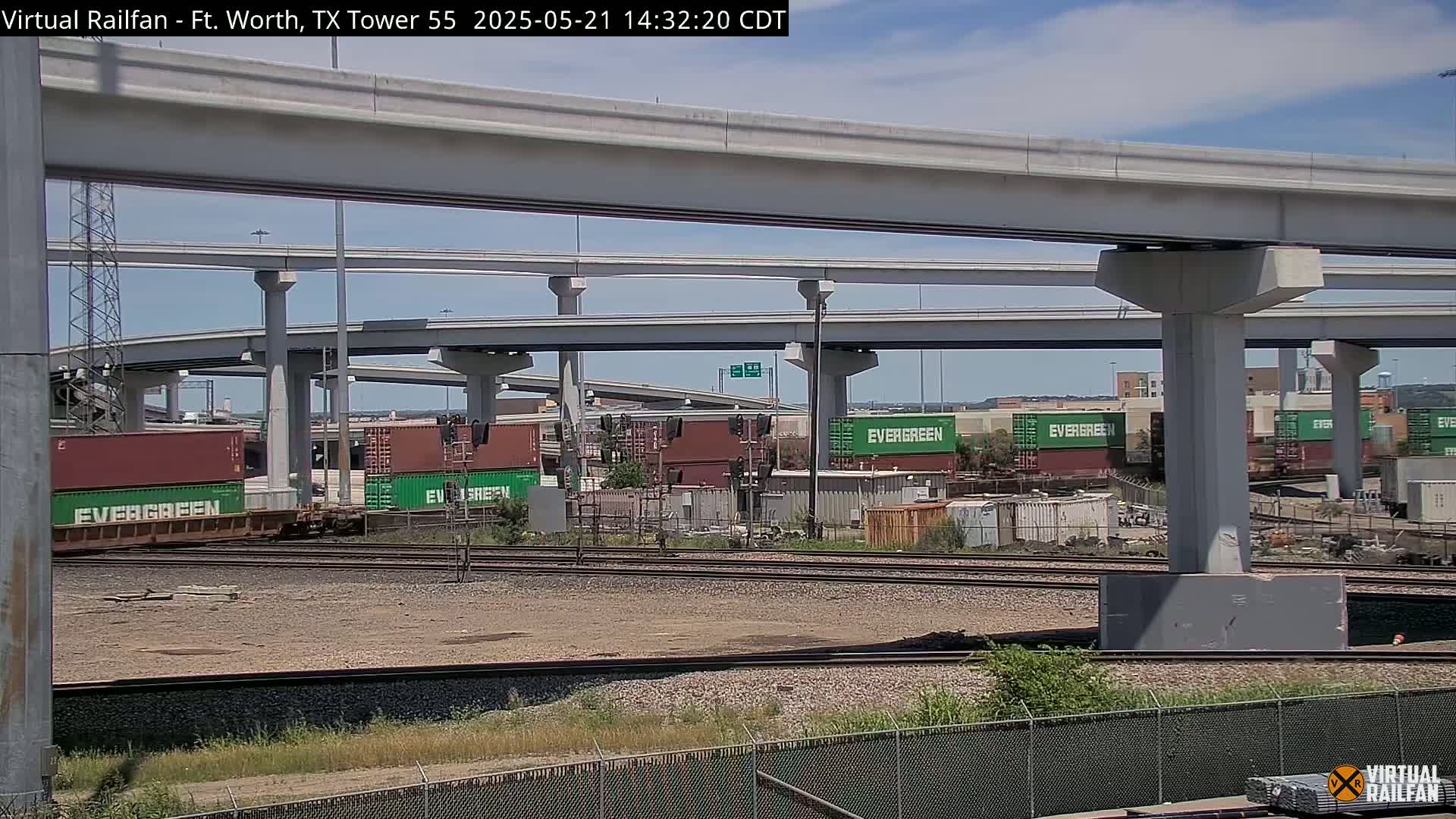 A train carrying Evergreen shipping containers sits below a multi-level highway overpass on a sunny day.