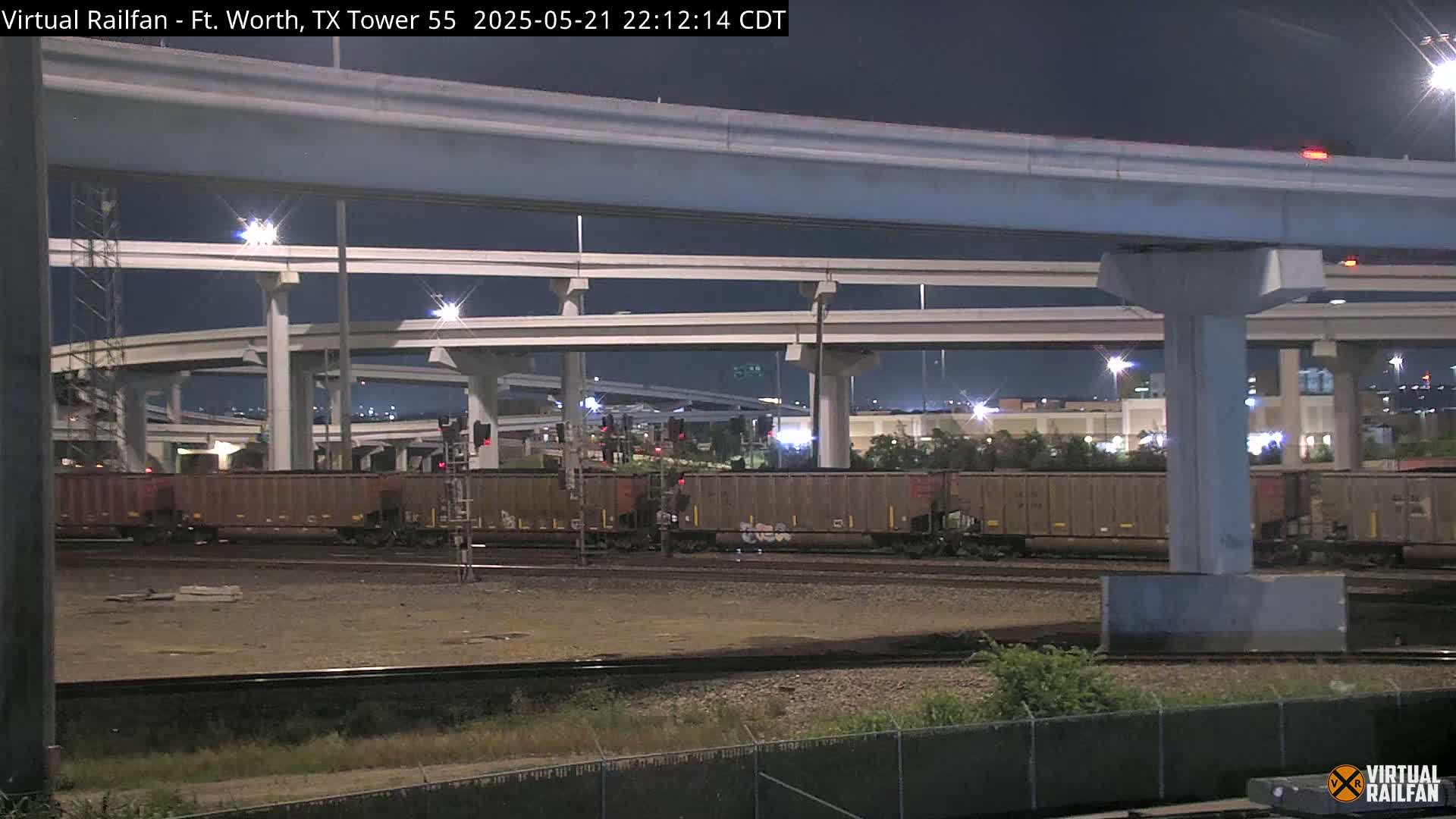 A nighttime view of a freight train stopped beneath an elevated highway interchange.