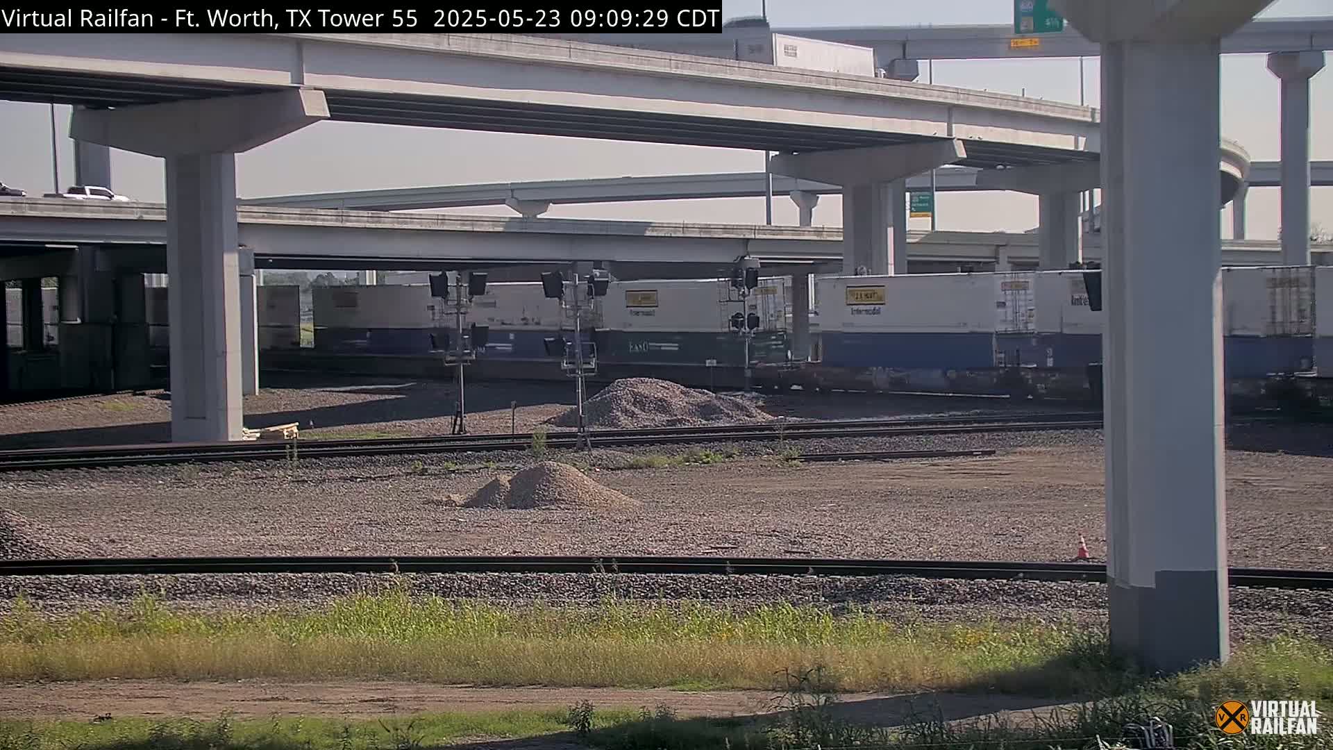Under a sunny sky, a freight train sits beneath a highway overpass near piles of gravel and railroad tracks.