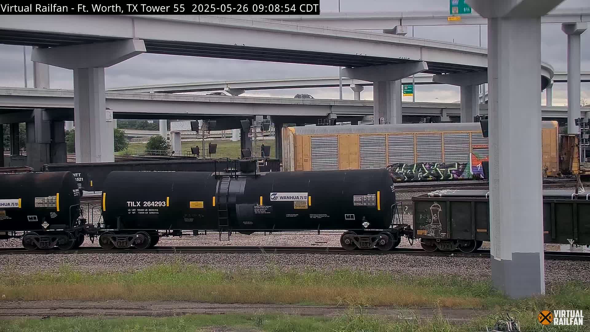 A train carrying tanker cars passes under an overpass on an overcast day.