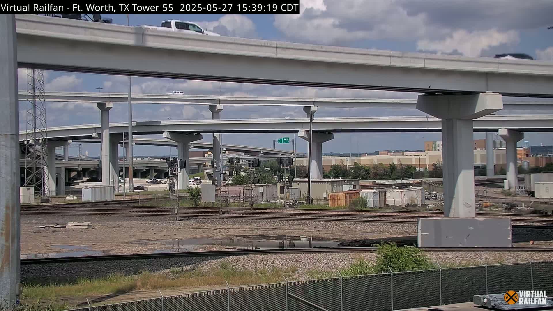 A partly cloudy sky is visible above a complex highway overpass system and adjacent railroad tracks, with some vehicles on the elevated roadways and a muddy area between the tracks and the highway.
