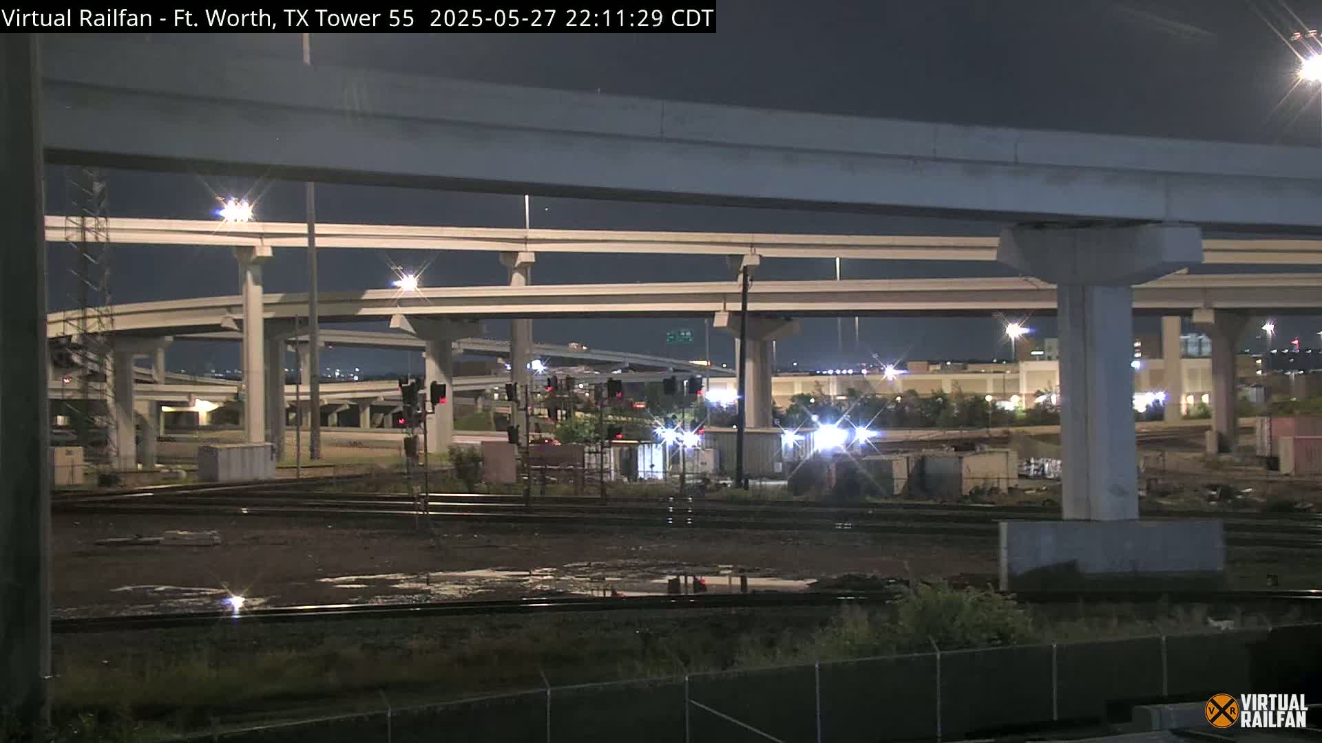 A nighttime view of a complex highway interchange over railroad tracks, with some puddles of water visible on the tracks.