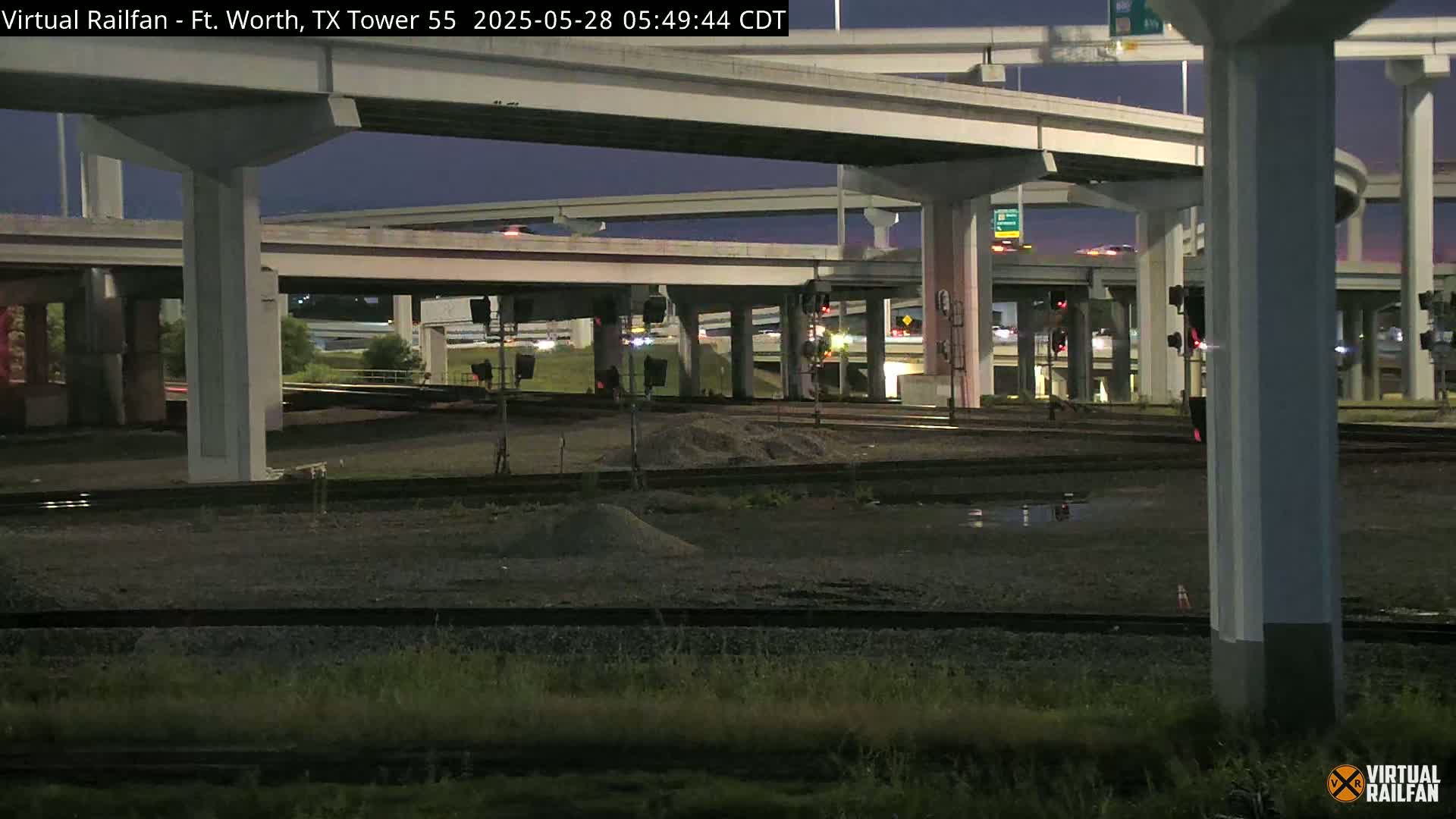 Under a twilight sky, a nighttime view shows a complex highway overpass intersecting with railroad tracks, with some puddles of water visible on the ground.