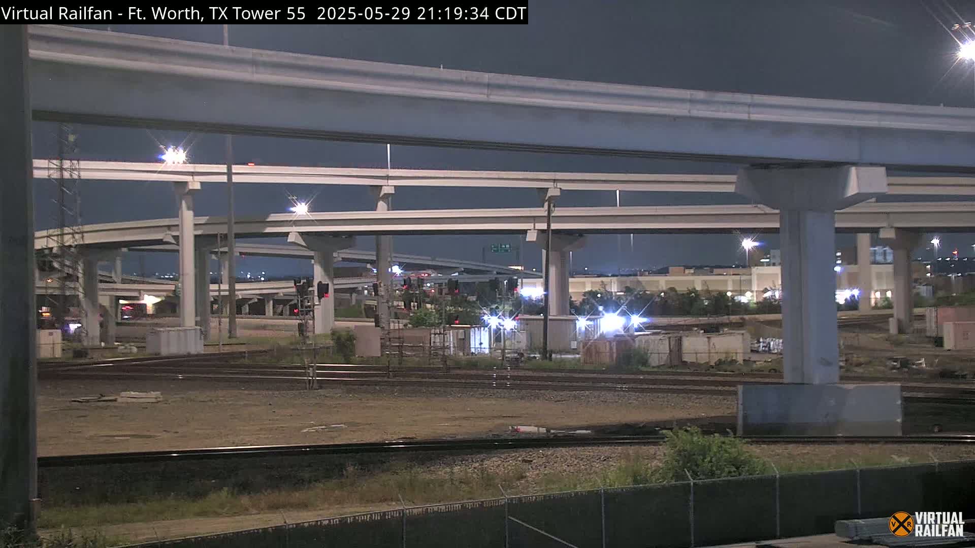 At night, under a dark sky, a complex highway overpass looms over railroad tracks and a construction or industrial area.