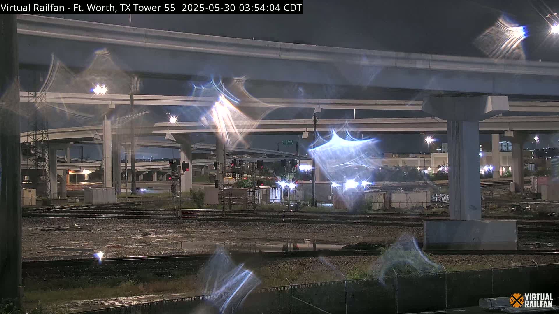 A nighttime view of a large highway overpass and train tracks, with wet ground and light reflections visible.