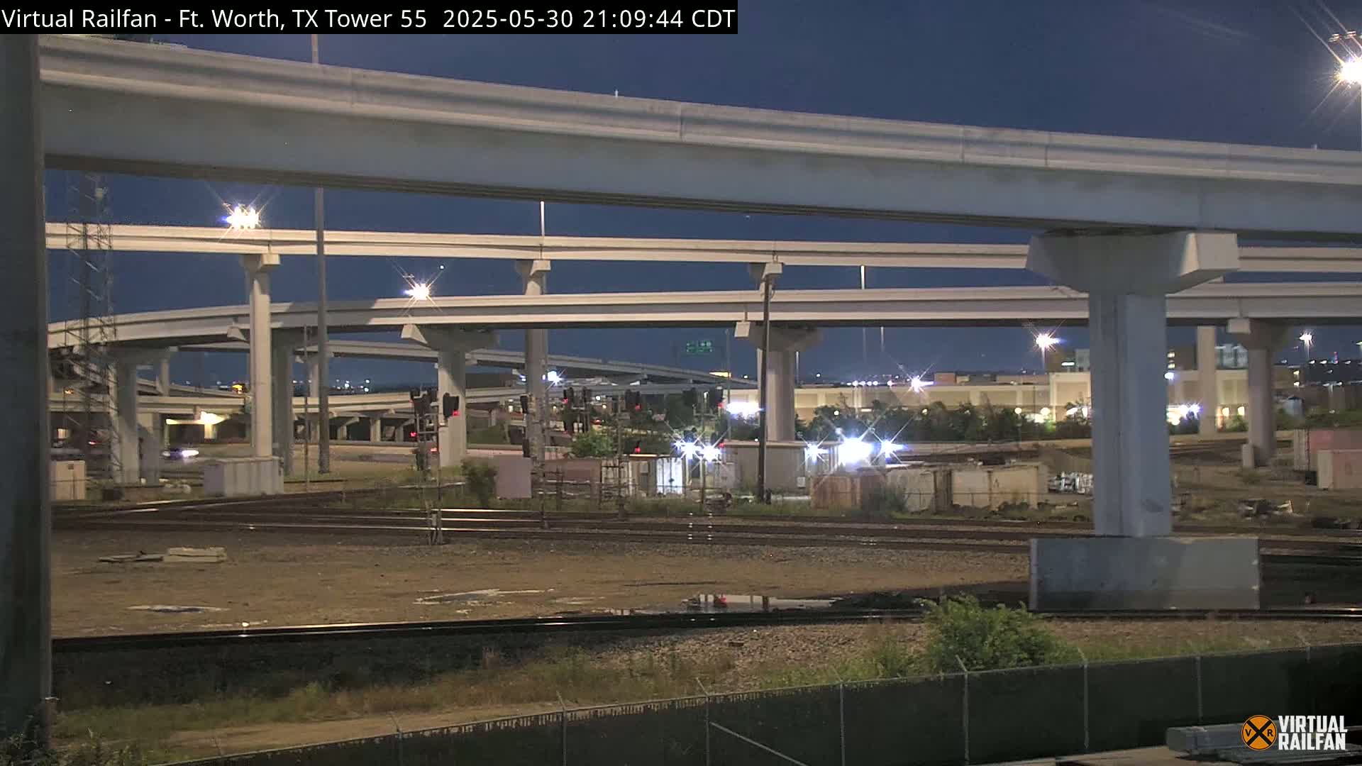 At night, under a clear sky, a complex highway overpass looms over railroad tracks and a sparsely vegetated area with some buildings and containers visible in the distance.