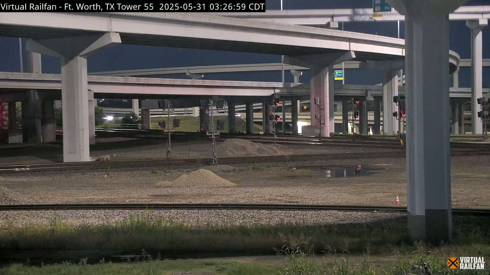 Under a dark night sky, railroad tracks and a large highway overpass are visible, with some small piles of gravel and a puddle of water in the foreground.