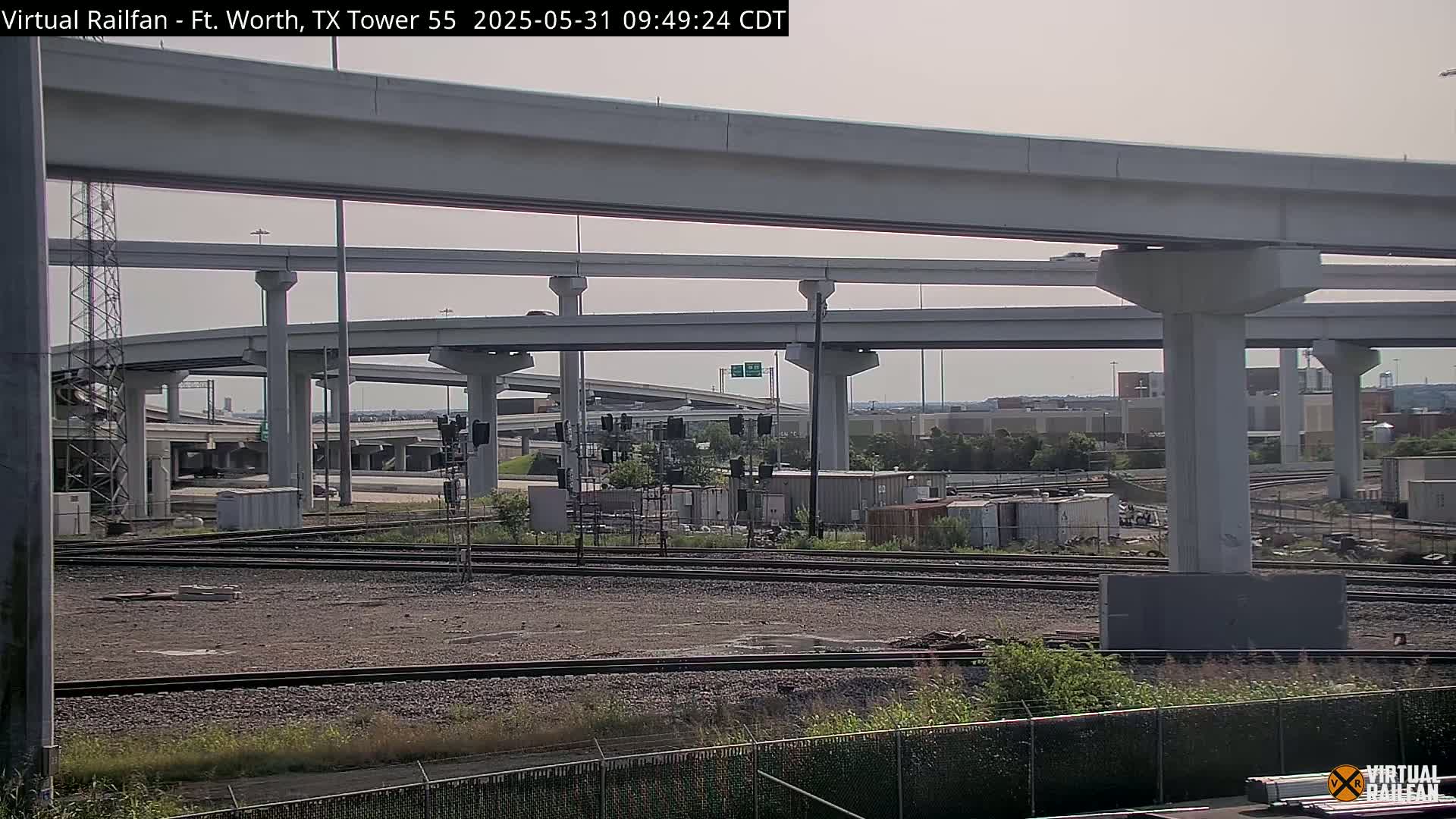 A view of a highway overpass with several layers of elevated roadways over railroad tracks and a fenced-in area, under a mostly clear sky.