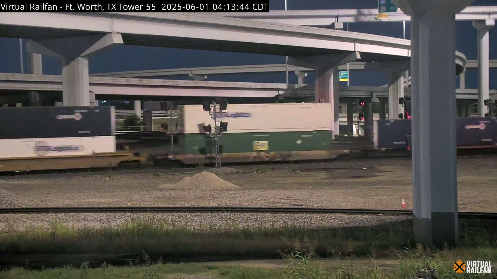 A freight train passes under a highway overpass at night.
