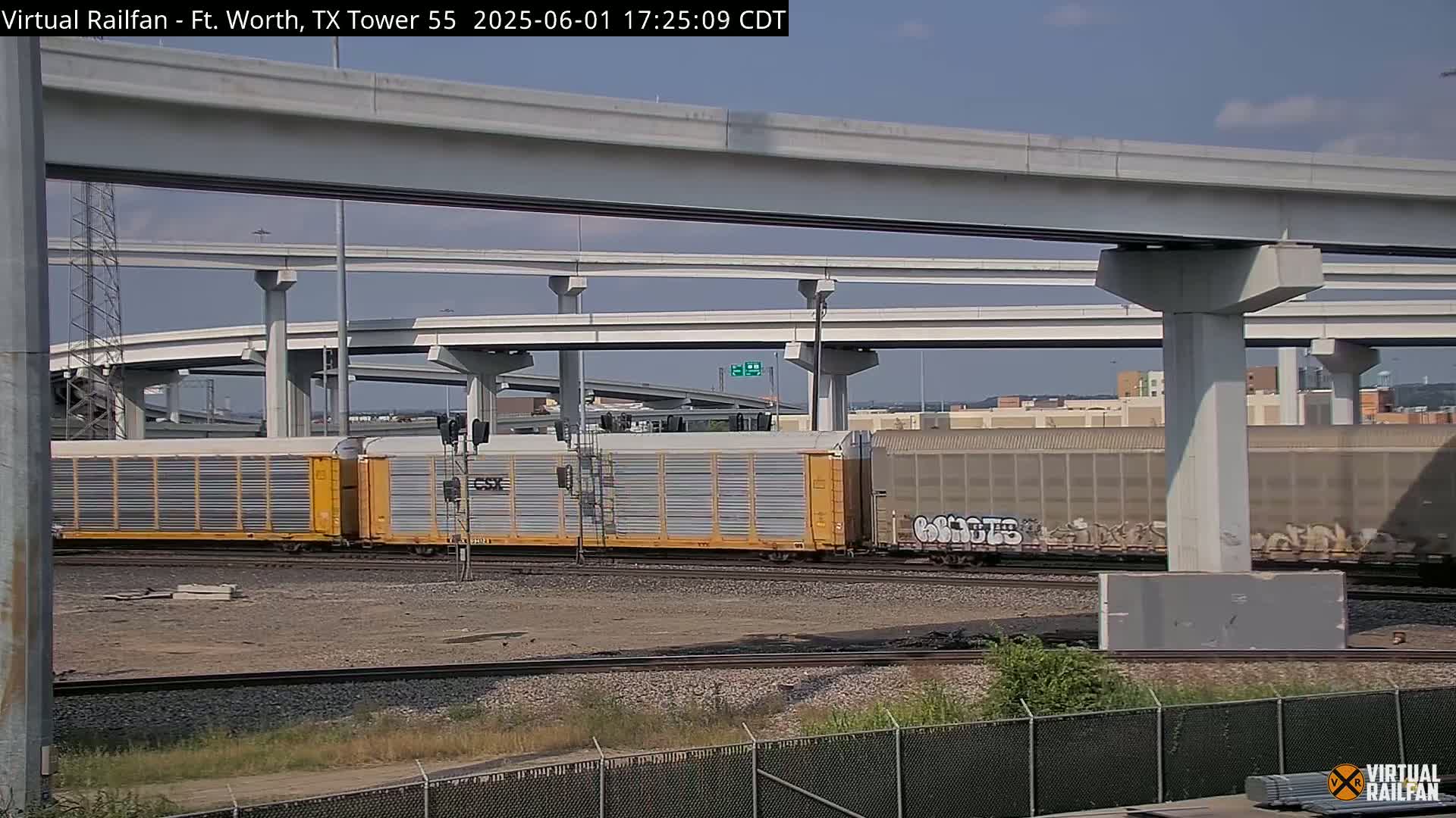 A freight train passes under a large highway overpass on a sunny day.