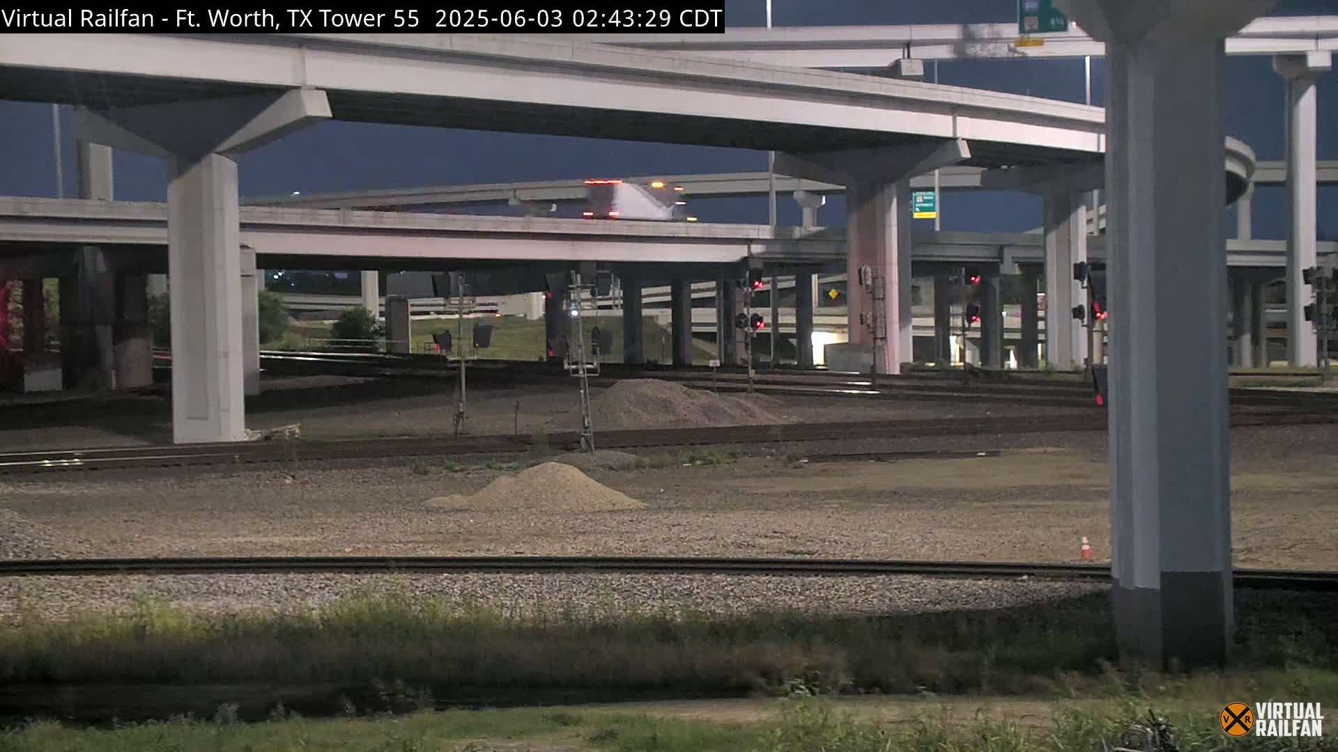 Under a dark night sky, a large highway overpass crosses over railroad tracks with small piles of gravel below.