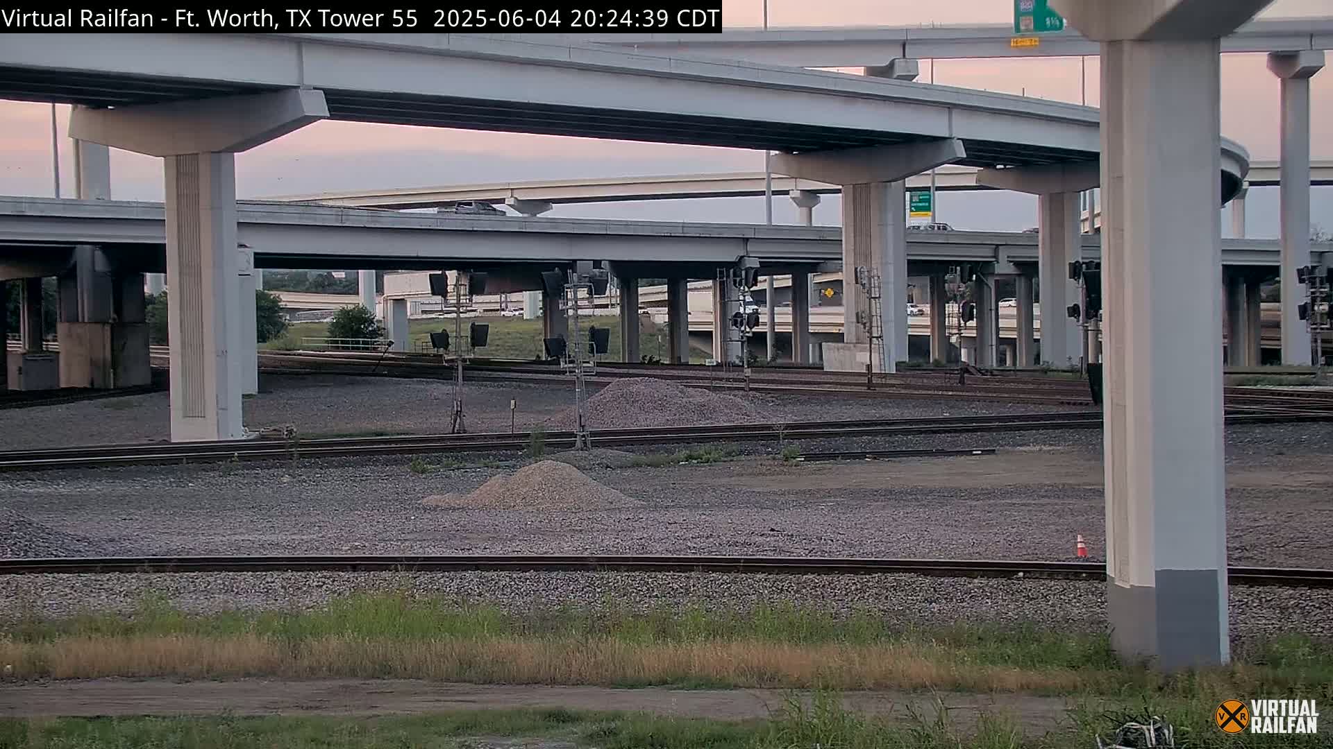 Under a twilight sky, railroad tracks and a highway overpass intersect, with small piles of gravel scattered between them.