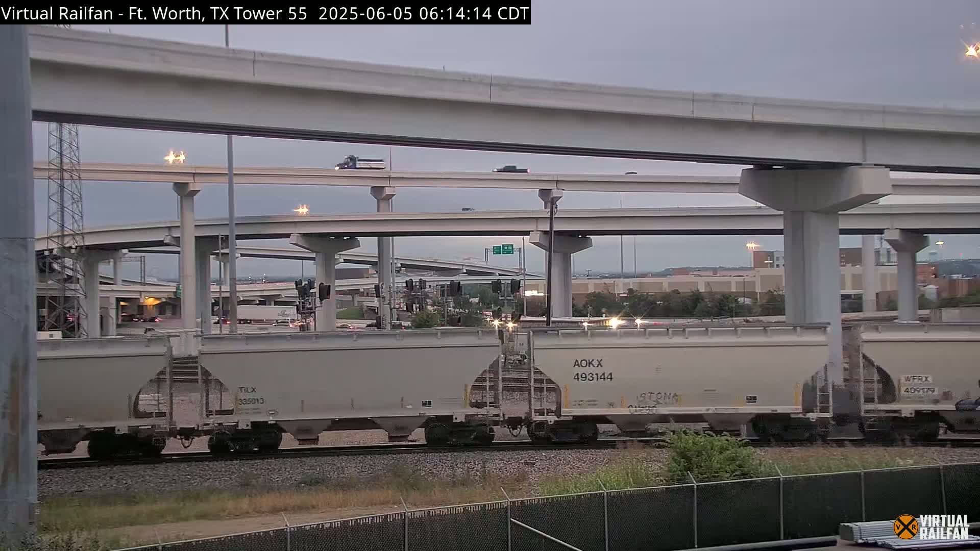 A freight train passes under a complex highway overpass system on an overcast day.