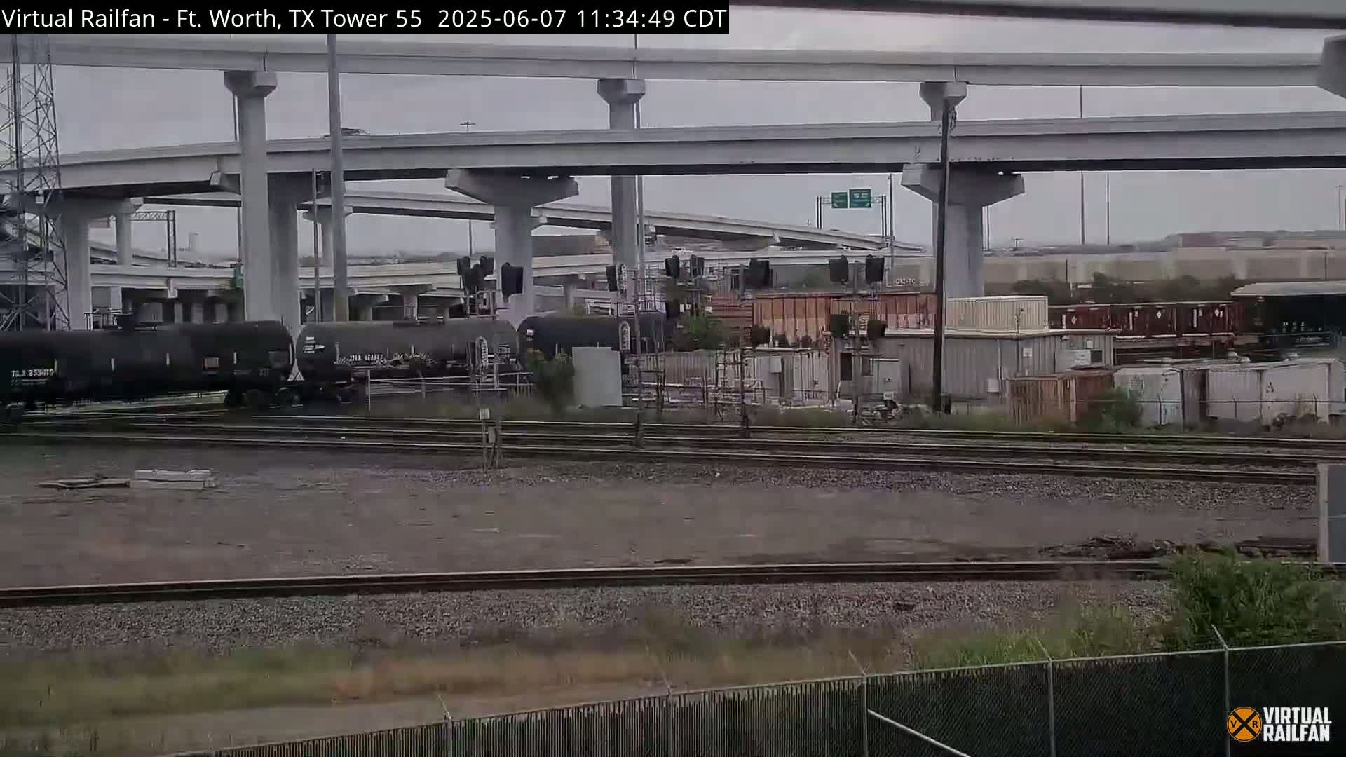 A railroad yard with several tanker cars sits beneath a large highway overpass on an overcast day.