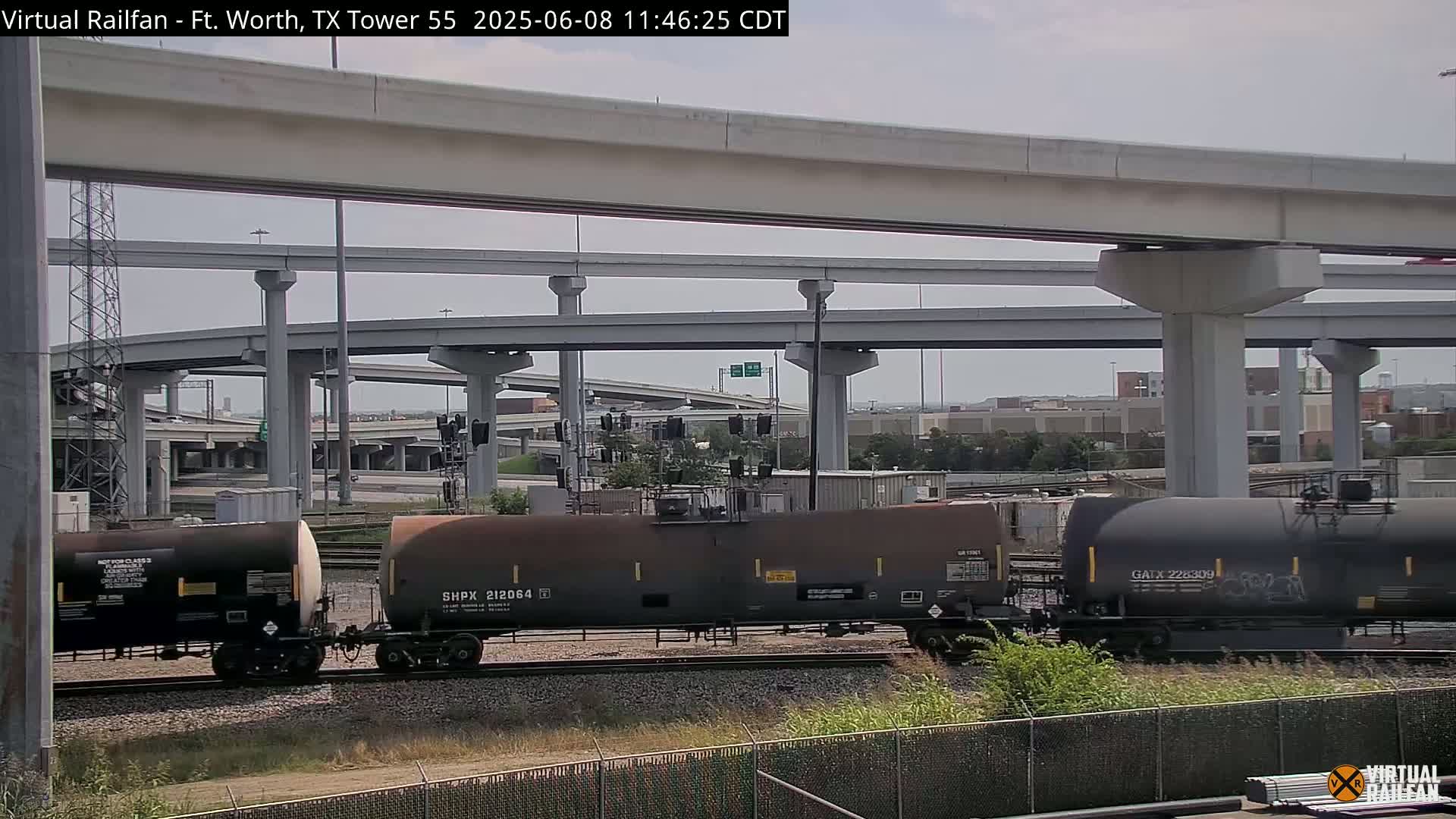 Under a partly cloudy sky, several railroad tank cars sit on tracks beneath a complex highway overpass.