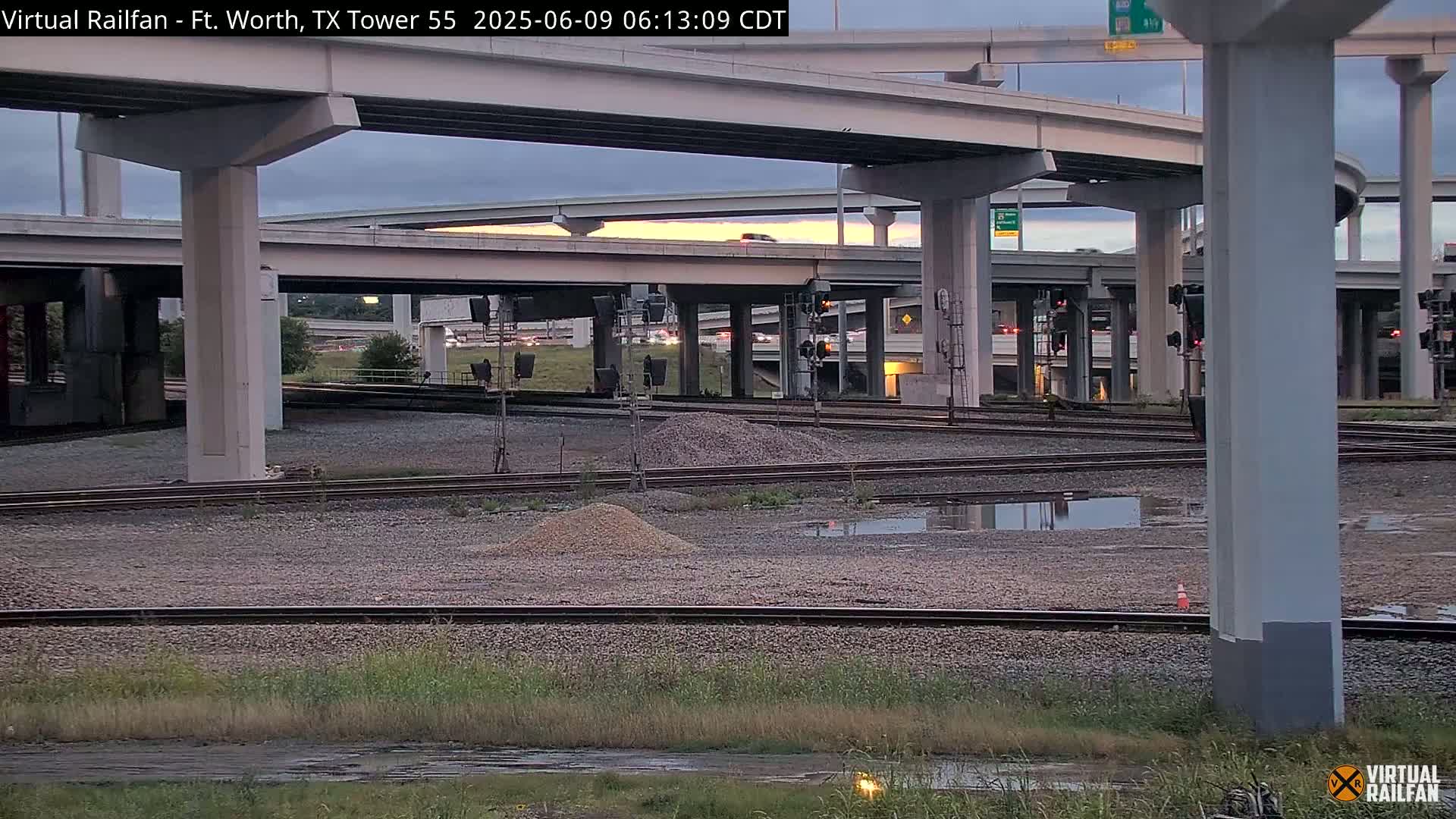 Under a partly cloudy twilight sky, railroad tracks and several highway overpasses intersect, with small piles of gravel and puddles of water visible on the ground below.