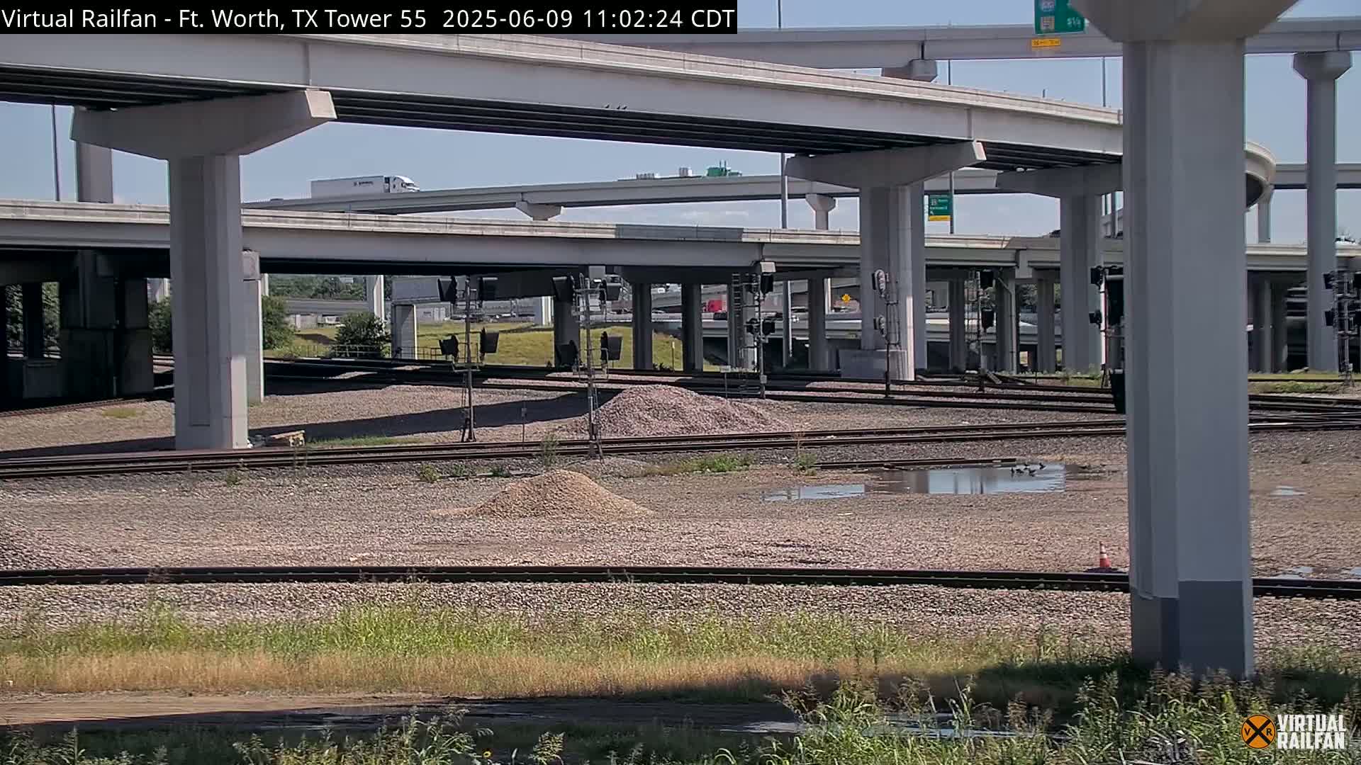 Under a sunny sky, railroad tracks intersect beneath a complex network of highway overpasses, with small piles of gravel and a puddle of water visible on the ground.