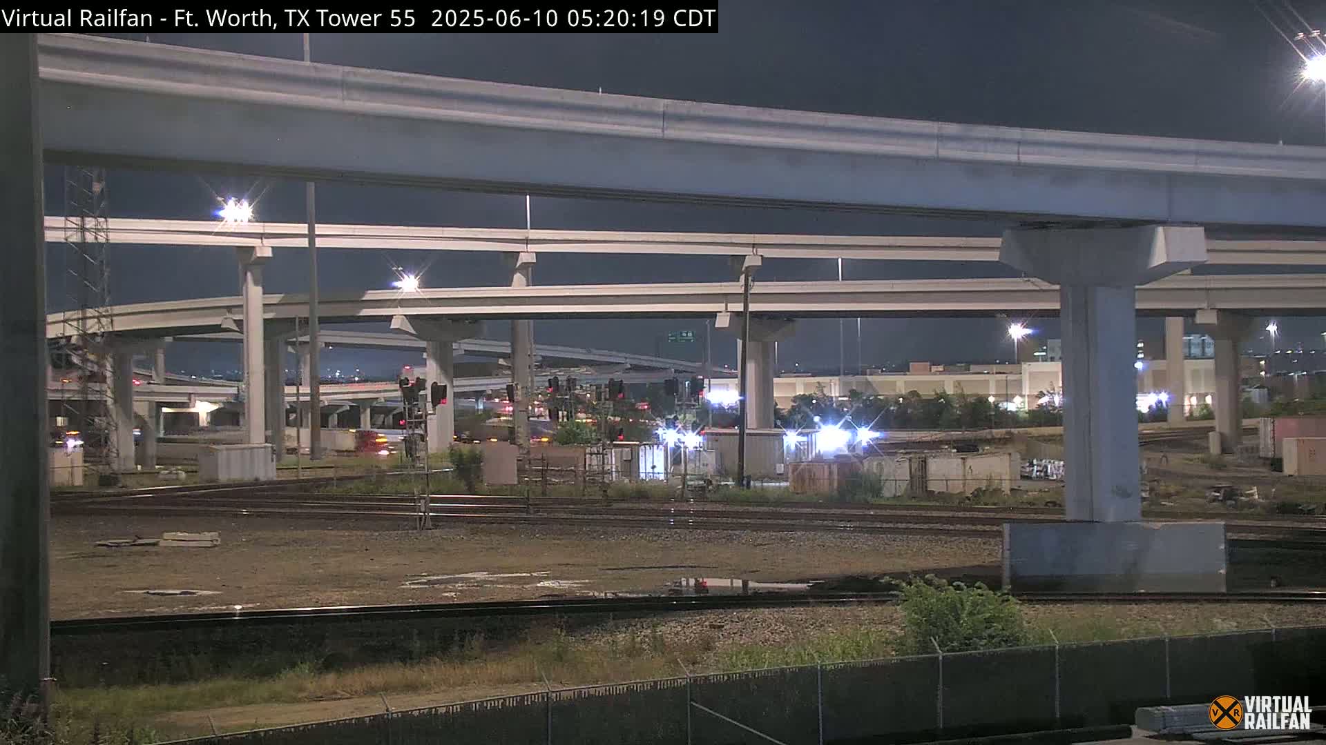 Under a dark night sky, a complex network of elevated roadways and train tracks intersect near a fenced-off area with some sparse vegetation.