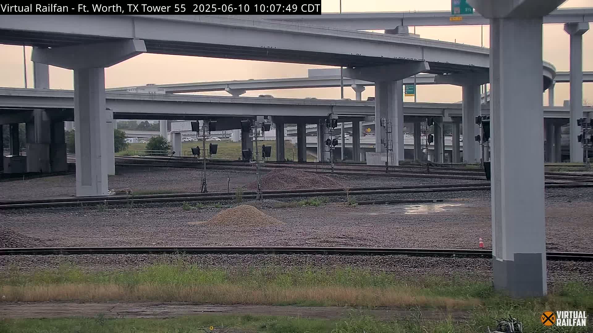 A view of railroad tracks beneath an overcast sky, surrounded by a complex network of highway overpasses and pillars.