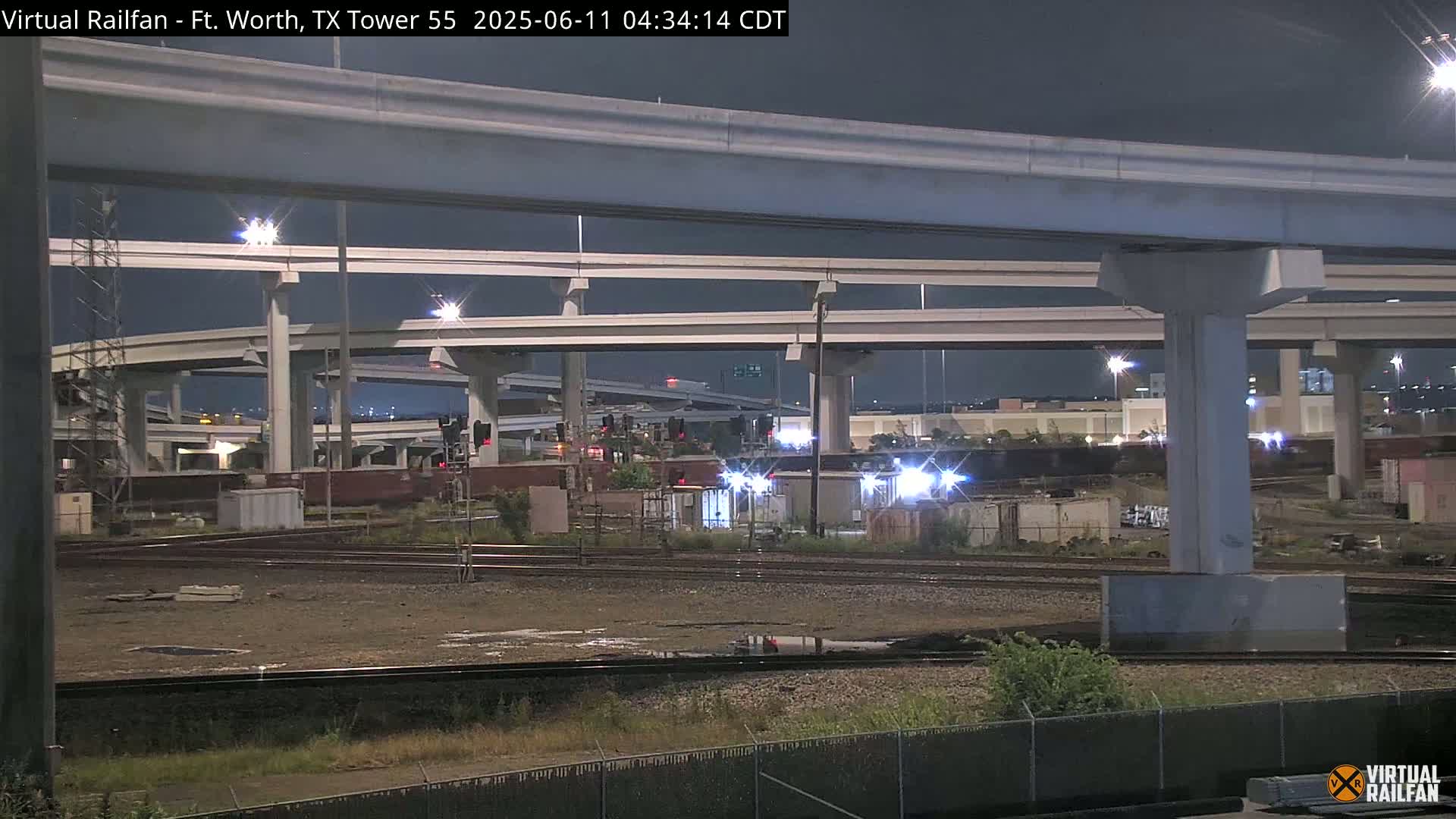 At night, under a dark sky, a nighttime view shows railroad tracks running under an elevated highway interchange.