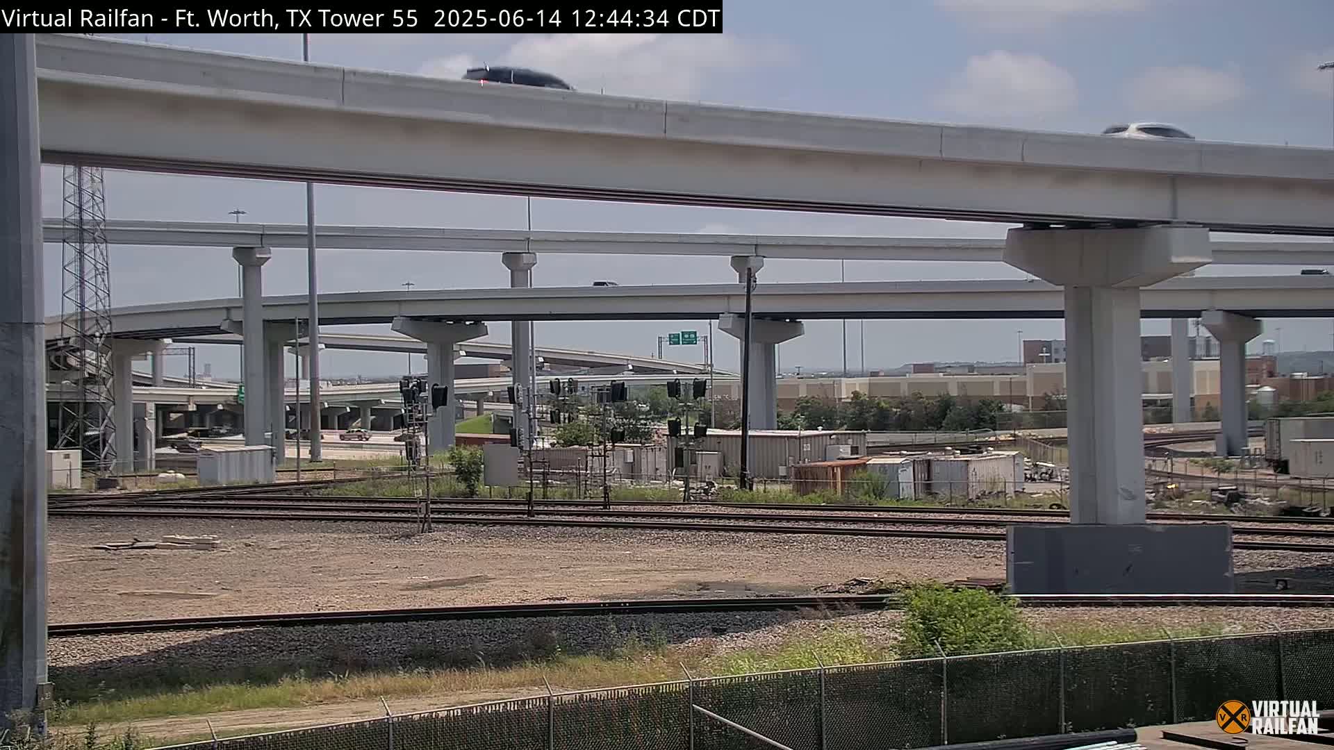 Under a partly cloudy sky, a complex highway interchange overpasses railroad tracks and a yard with shipping containers.