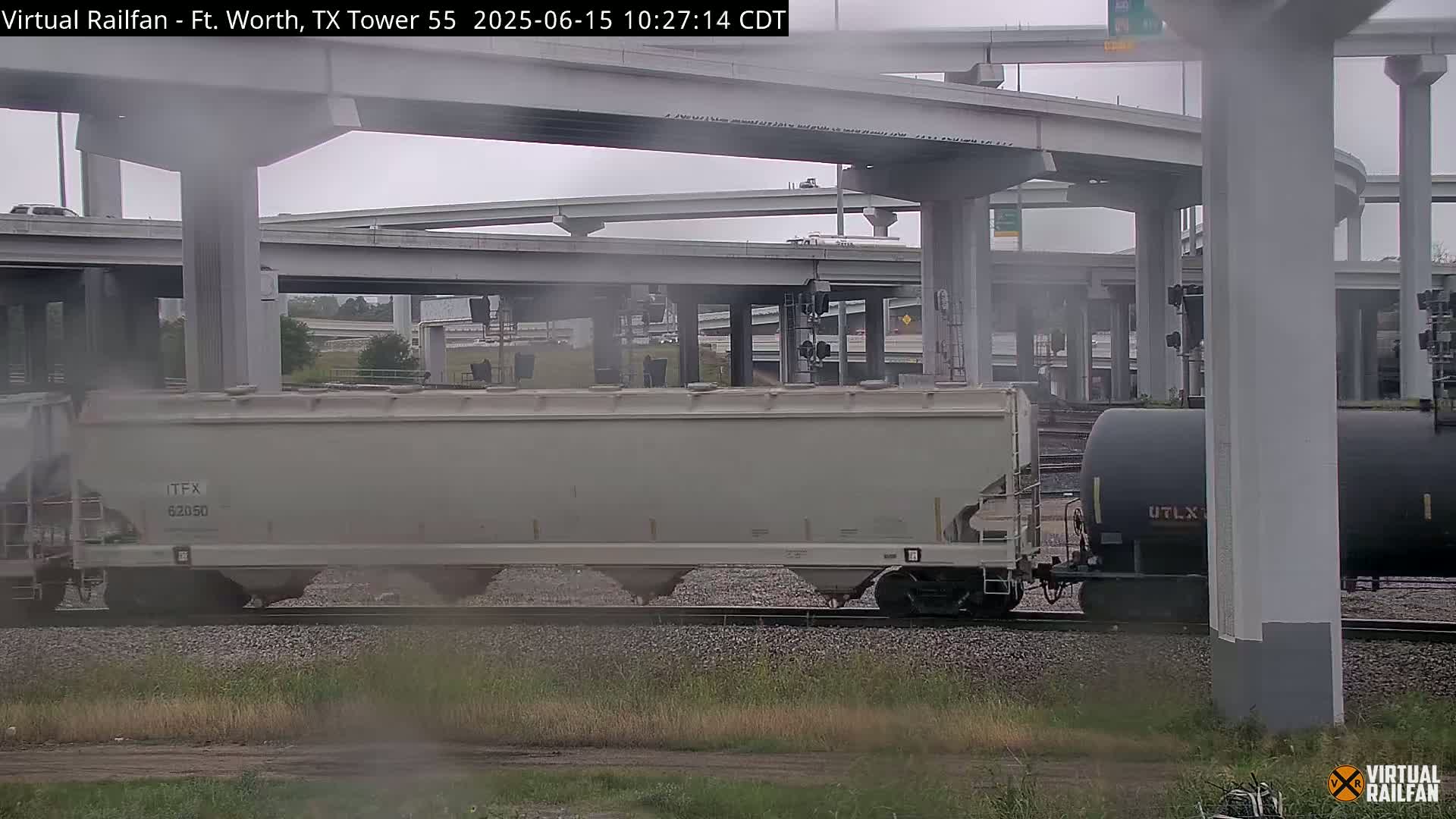 A freight train passes under a large overpass on an overcast day.