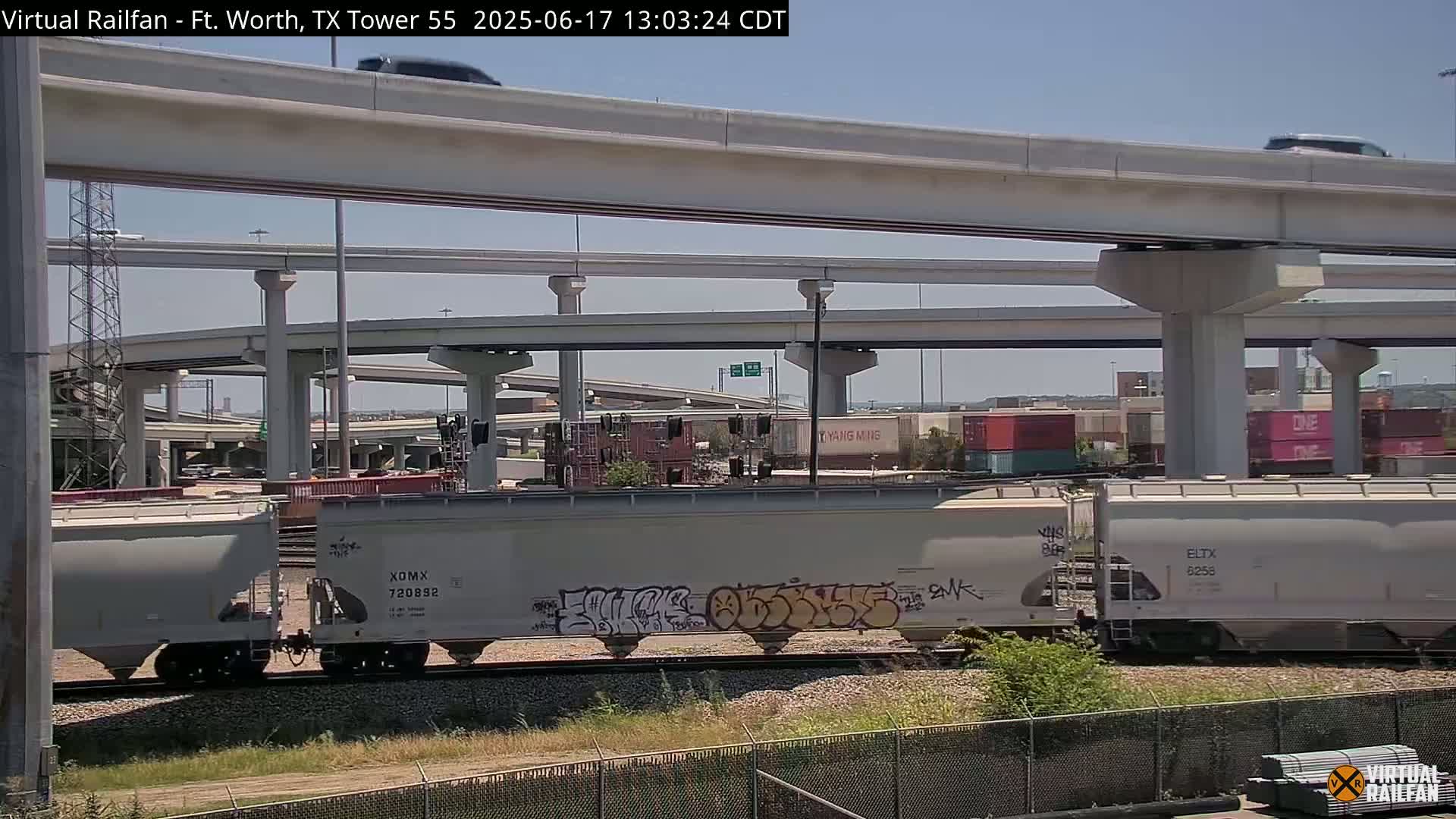 A freight train with graffiti on its cars passes beneath a complex highway overpass on a sunny day.