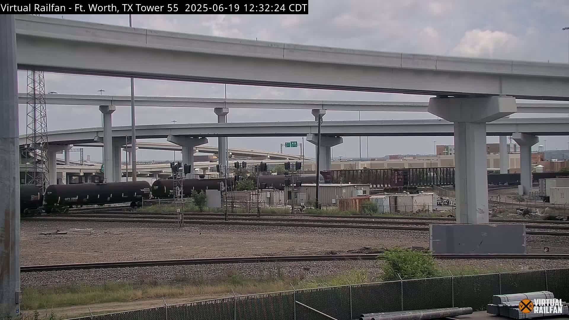 Under an overcast sky, several rail cars sit on a track below a complex overpass system.
