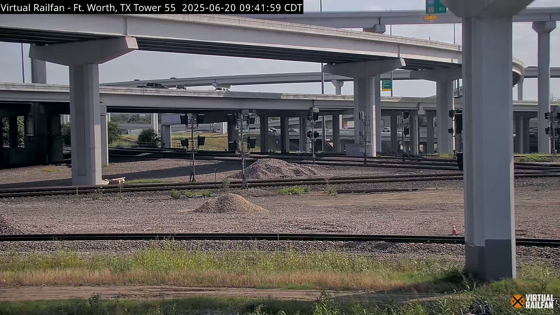 A view of a highway overpass and railway tracks under mostly sunny skies, with piles of gravel visible near the tracks.