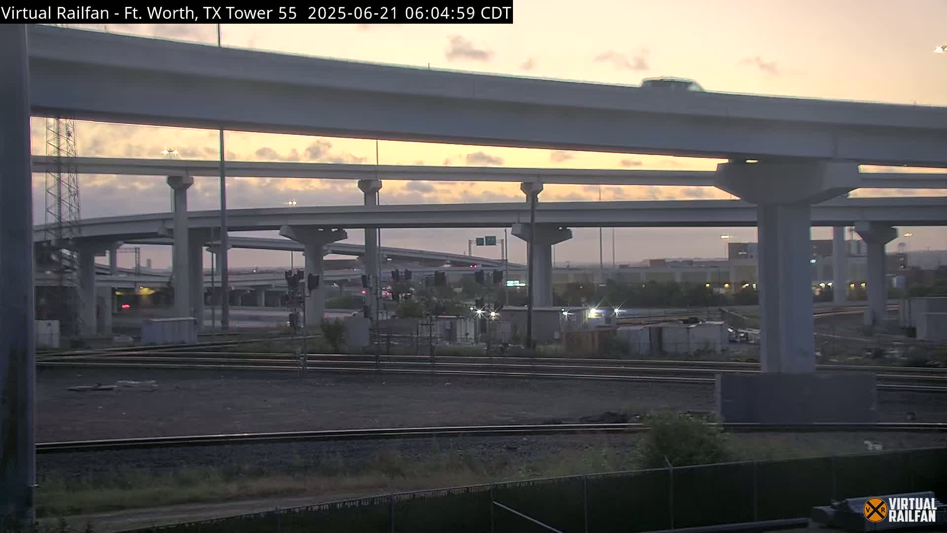 Under a twilight sky, an elevated highway interchange spans over railroad tracks and a flat, sparsely vegetated area.