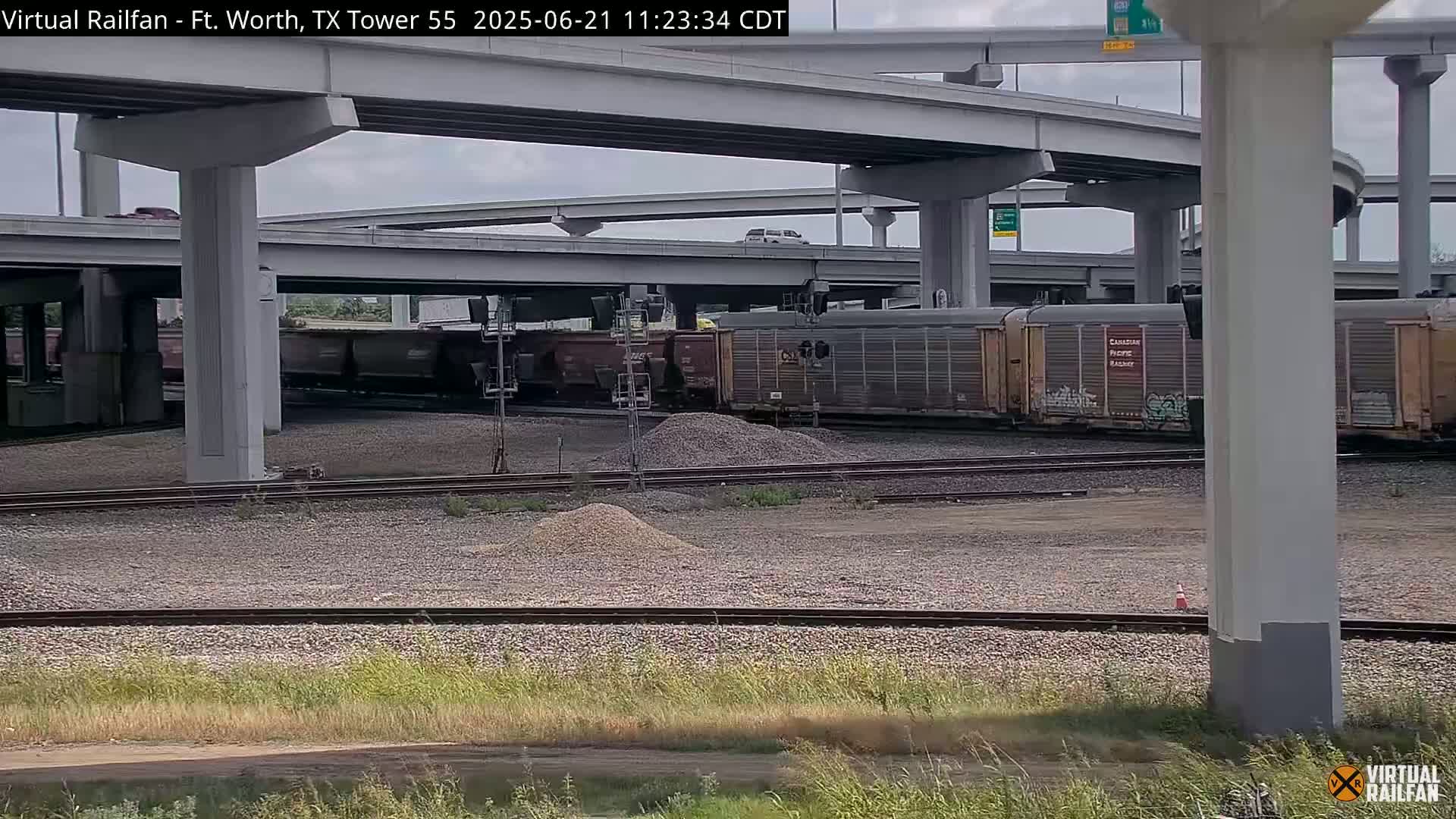 A freight train sits on railway tracks under a highway overpass on an overcast day.