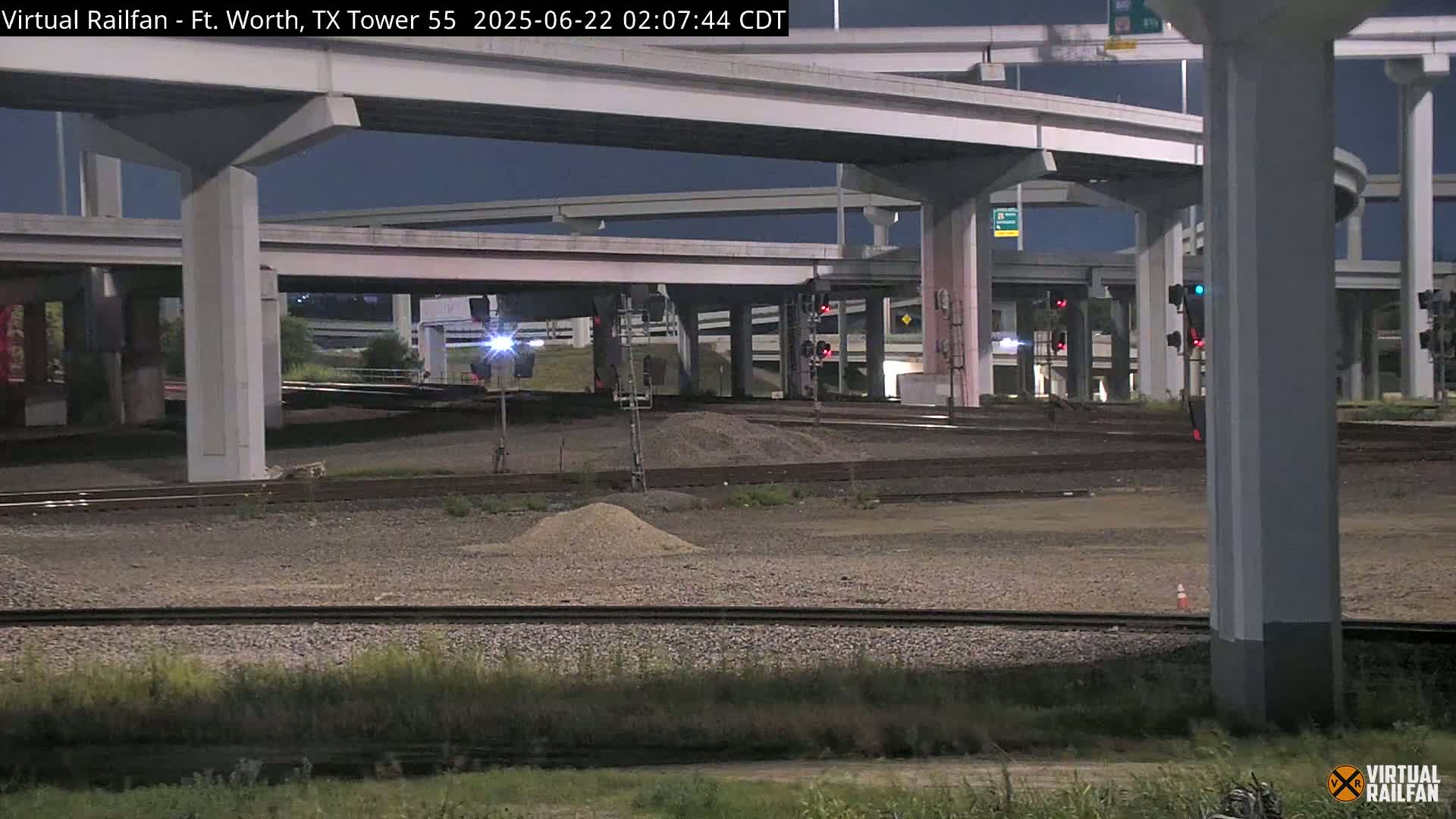 Under a nighttime sky, railroad tracks run beneath a complex overpass system, with small piles of gravel scattered on the ground.