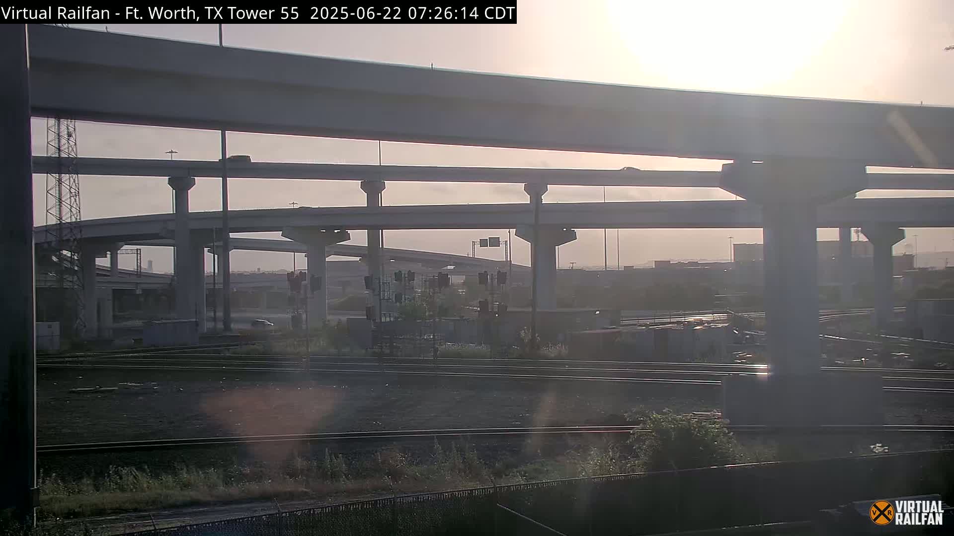 A hazy sunrise illuminates a complex highway overpass system above railroad tracks and a largely empty industrial area.