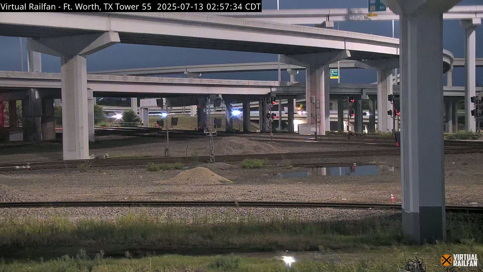 Under a dark night sky, a complex highway interchange overpasses railroad tracks, with some puddles present in the area below.