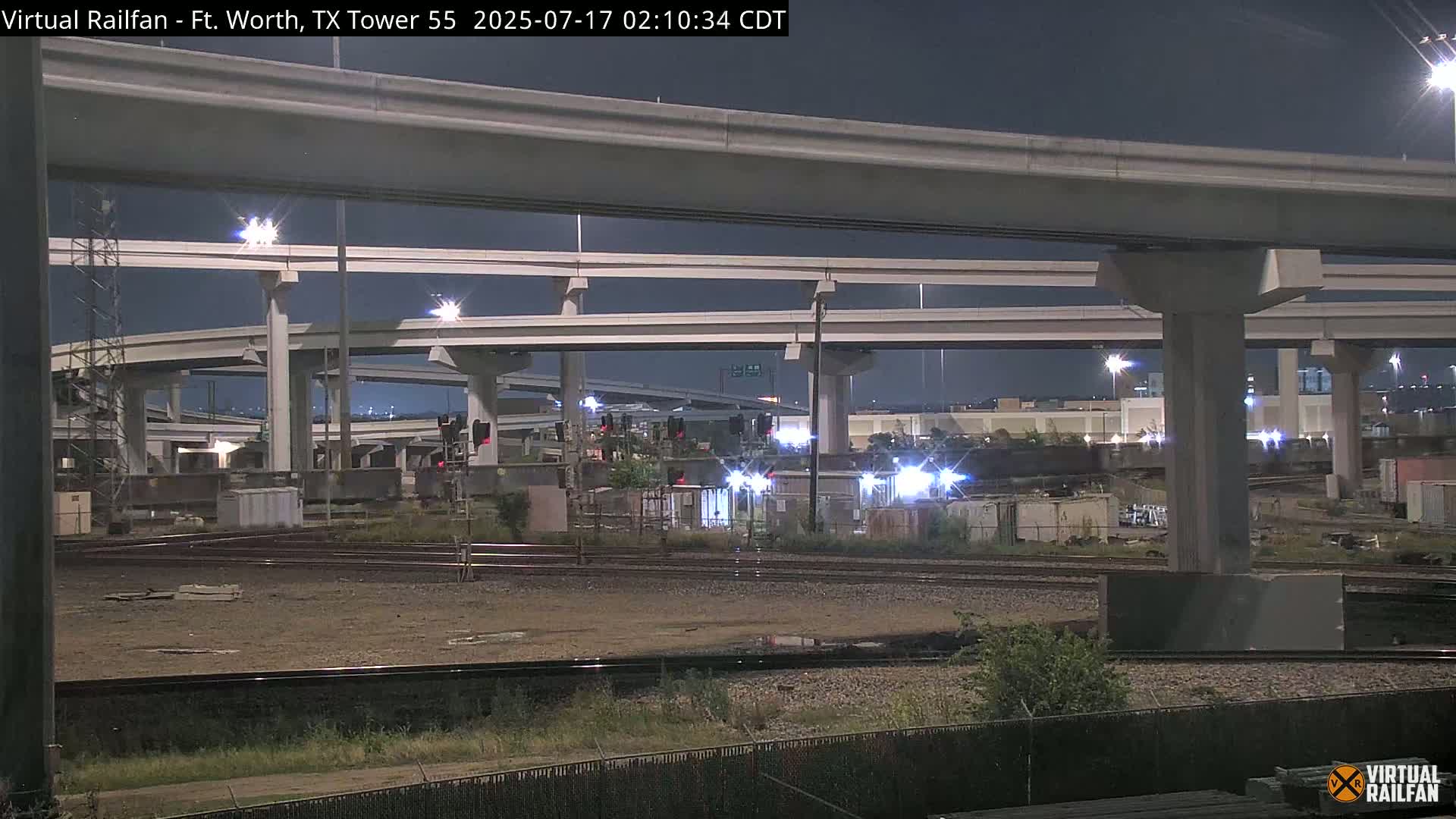 At night, a complex highway overpass looms over railroad tracks and a mostly empty industrial area.
