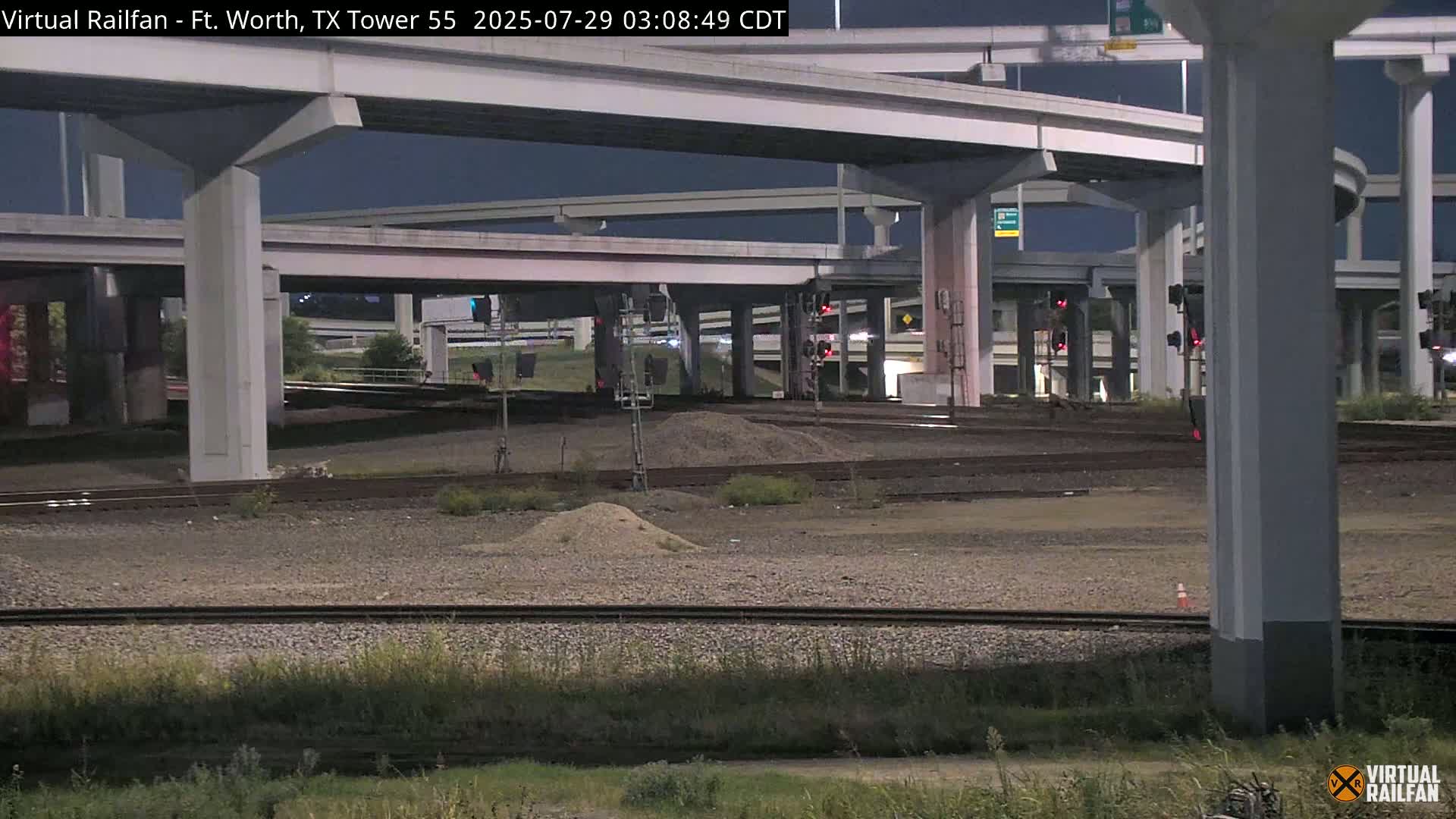 At night, railroad tracks intersect beneath a complex overpass system of elevated roadways.