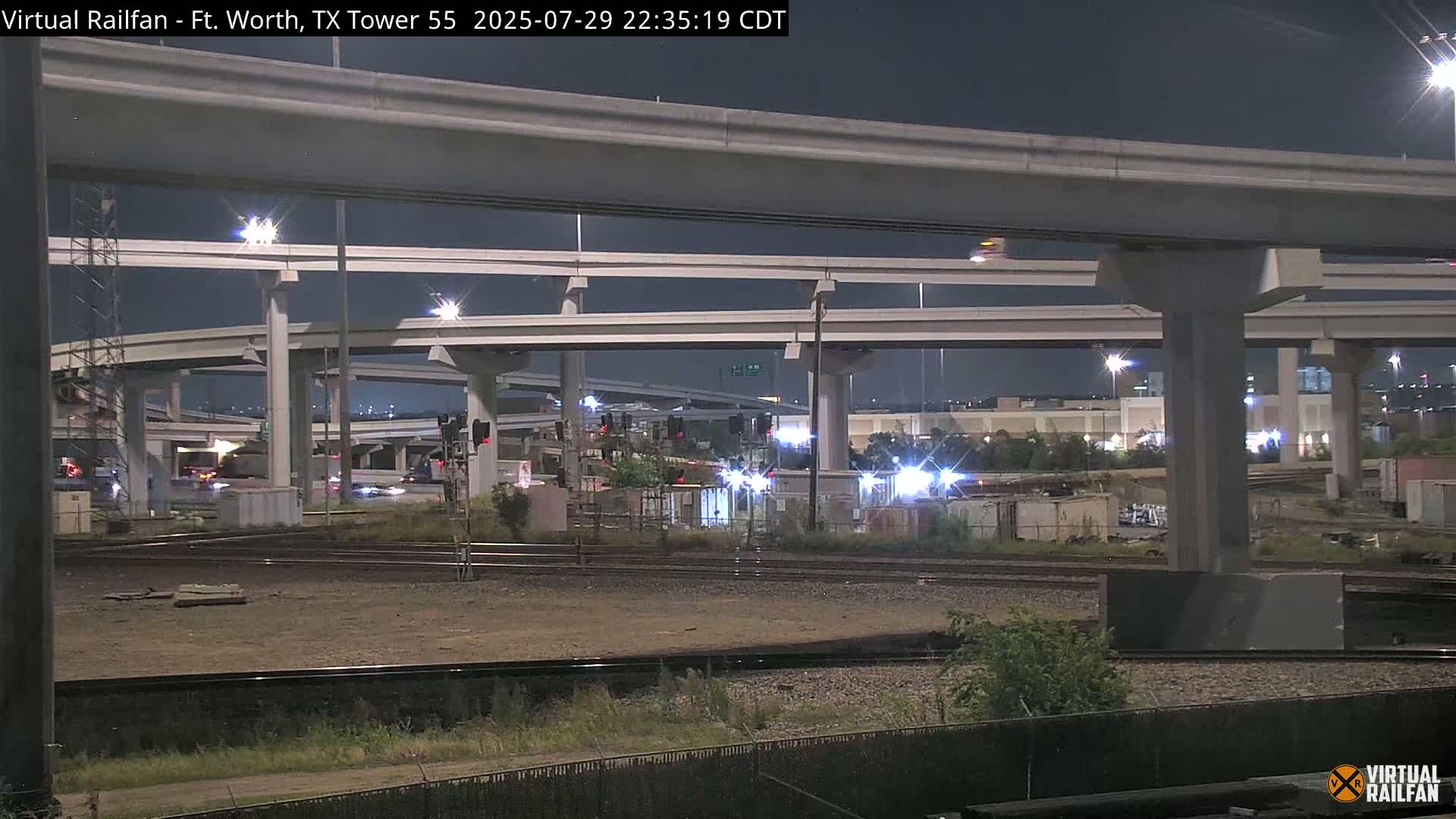 At night, railroad tracks intersect beneath a complex overpass system of elevated roadways.